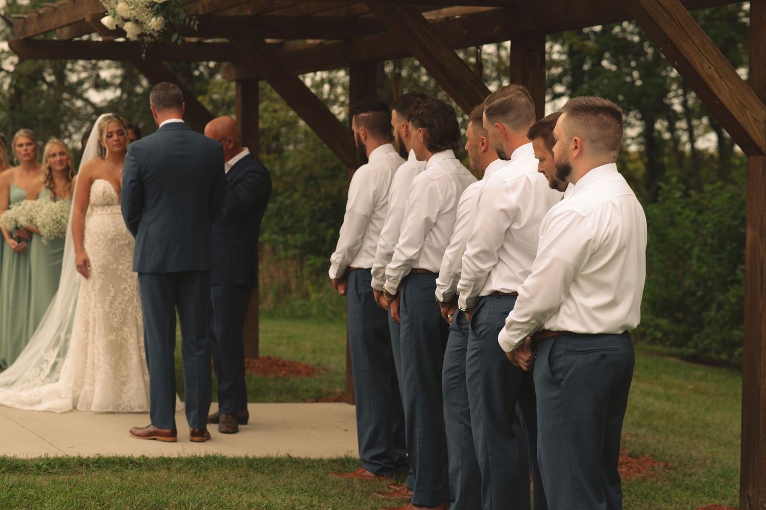 Wedding ceremony participants gather under wooden beams in an outdoor setting.