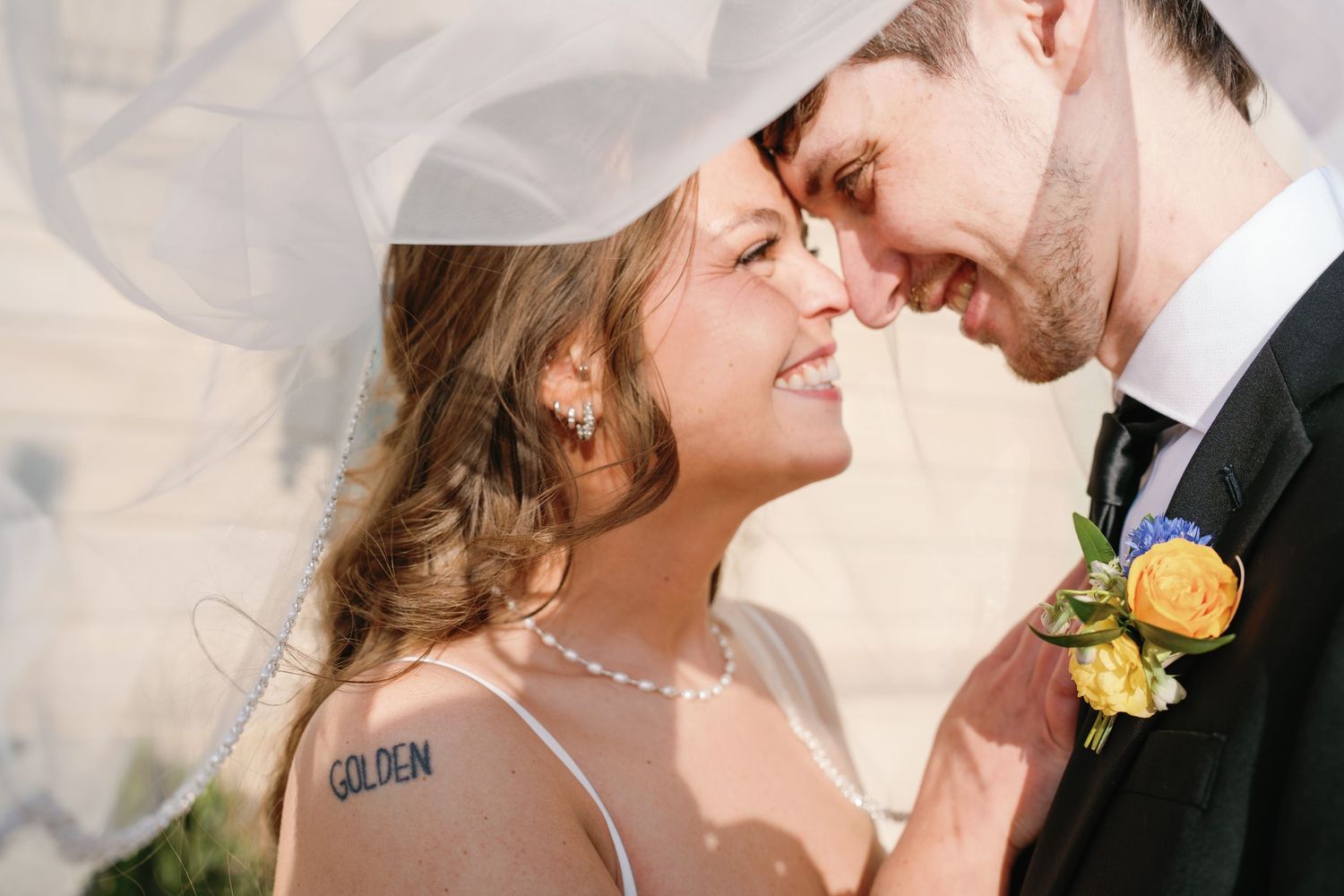 Bride and groom touching foreheads under veil, bride with GOLDEN tattoo wearing pearl necklace, groom in suit with boutonniere.