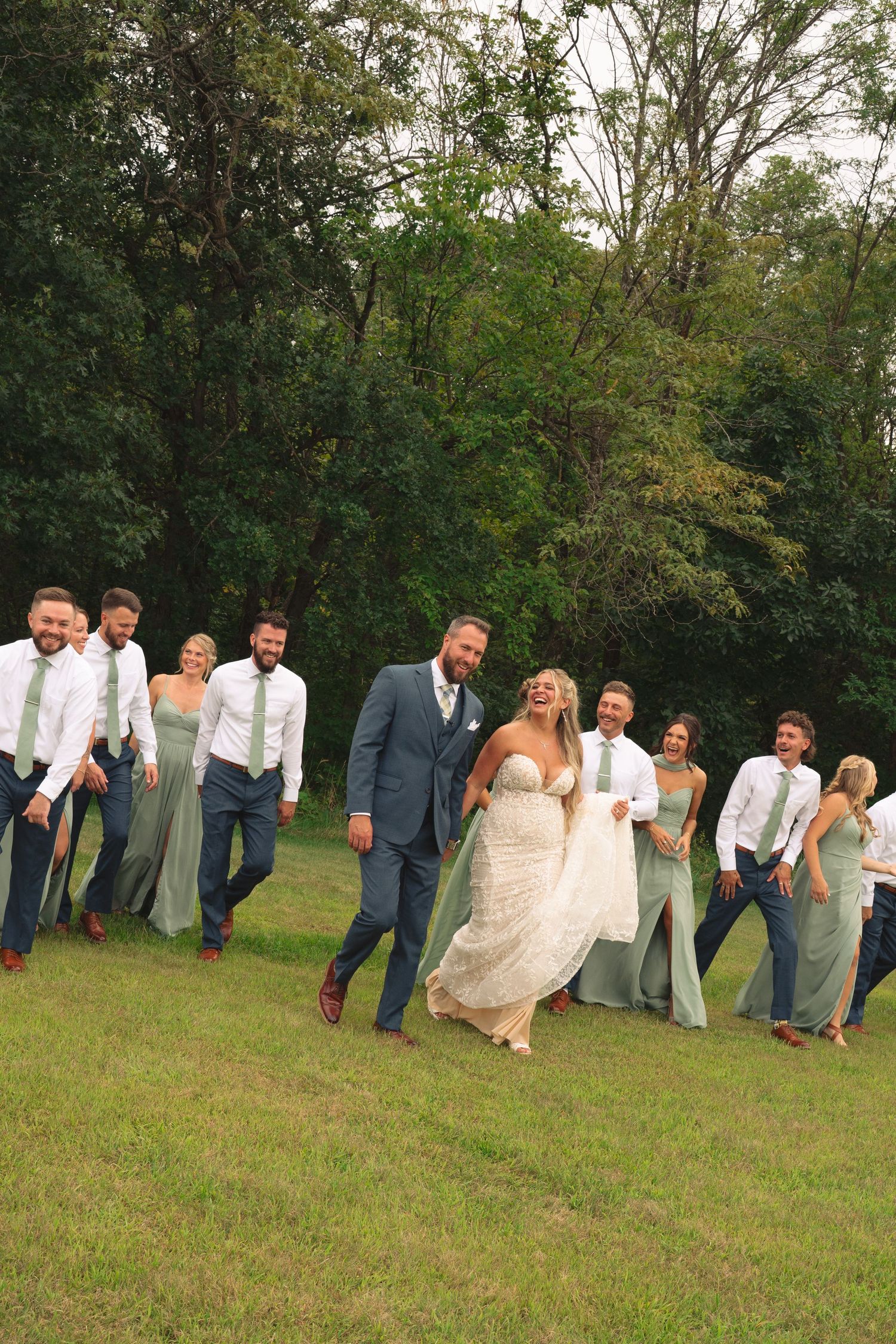 Wedding party poses playfully on grassy field with rustic backdrop.