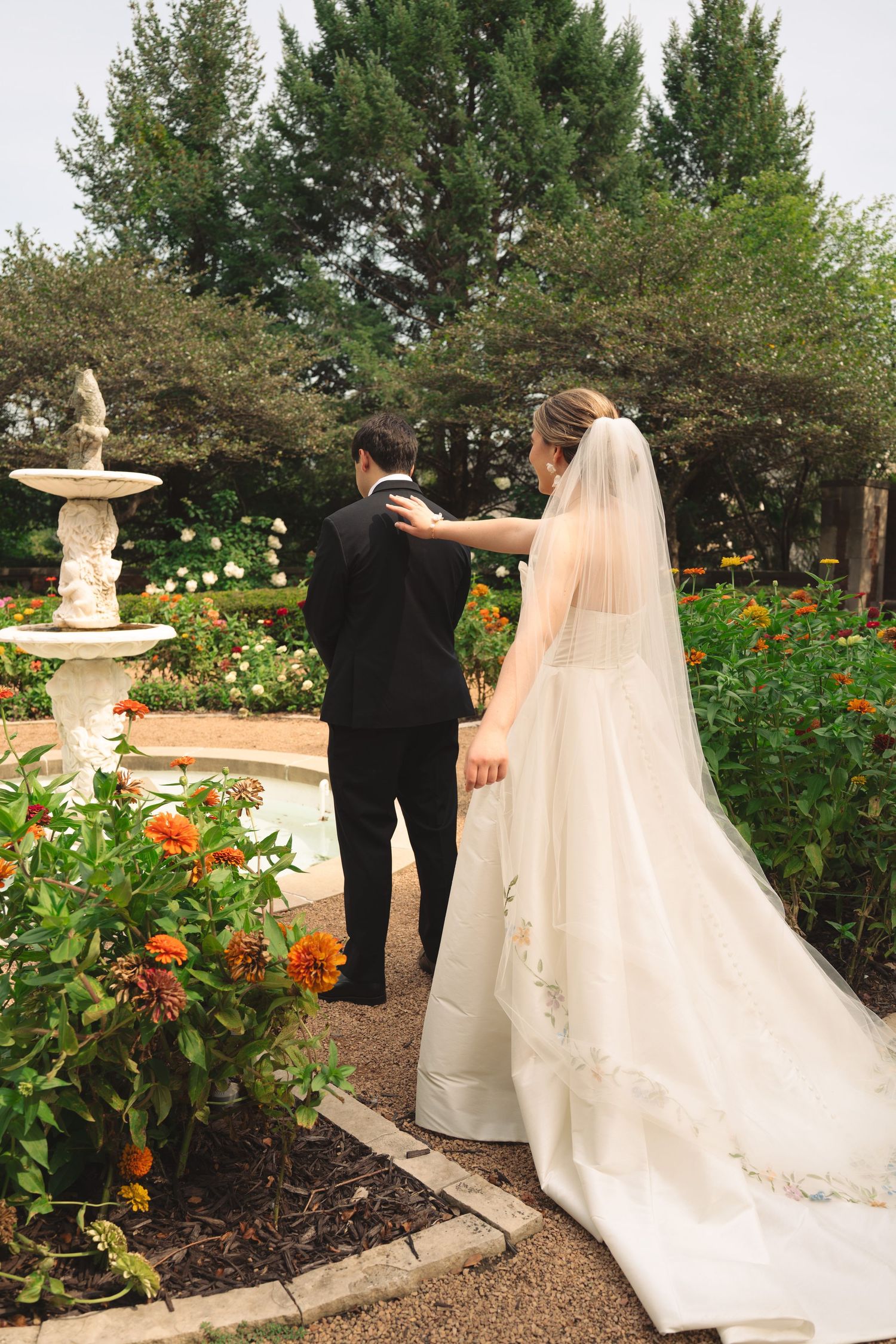 Wedding couple walking through manicured garden path with decorative stone fountain.