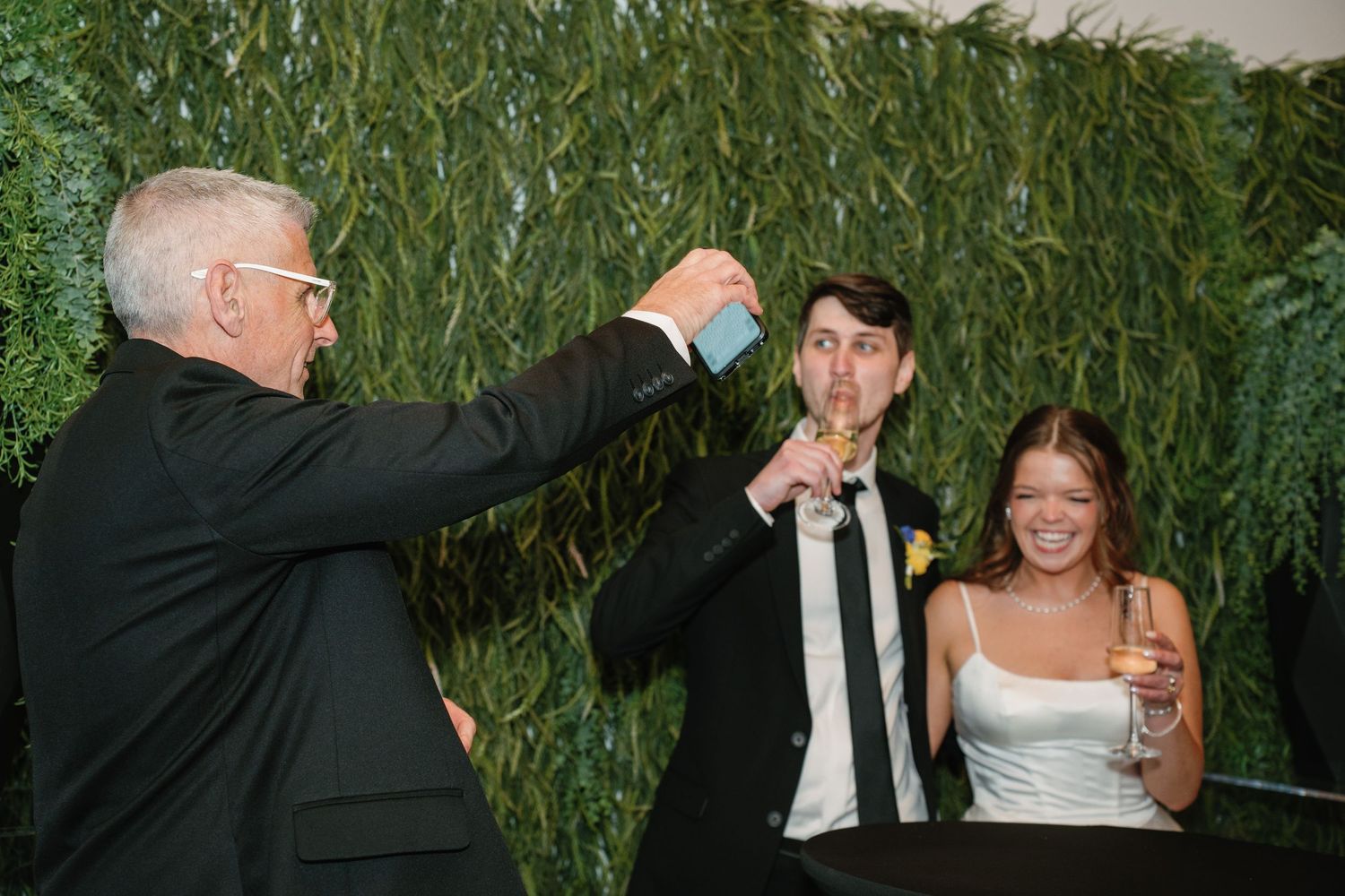 Older man in black suit raises glass in toast with bride and groom at wedding reception near green wall.