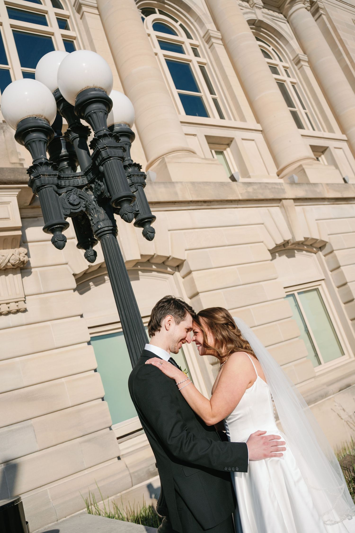 Bride and groom sharing a romantic moment under a street lamp outside a grand stone building on their wedding day.