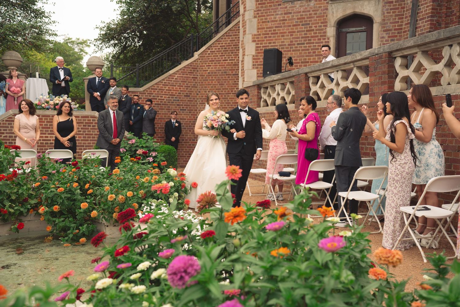 Wedding guests seated in garden setting with colorful flowers and brick architecture.