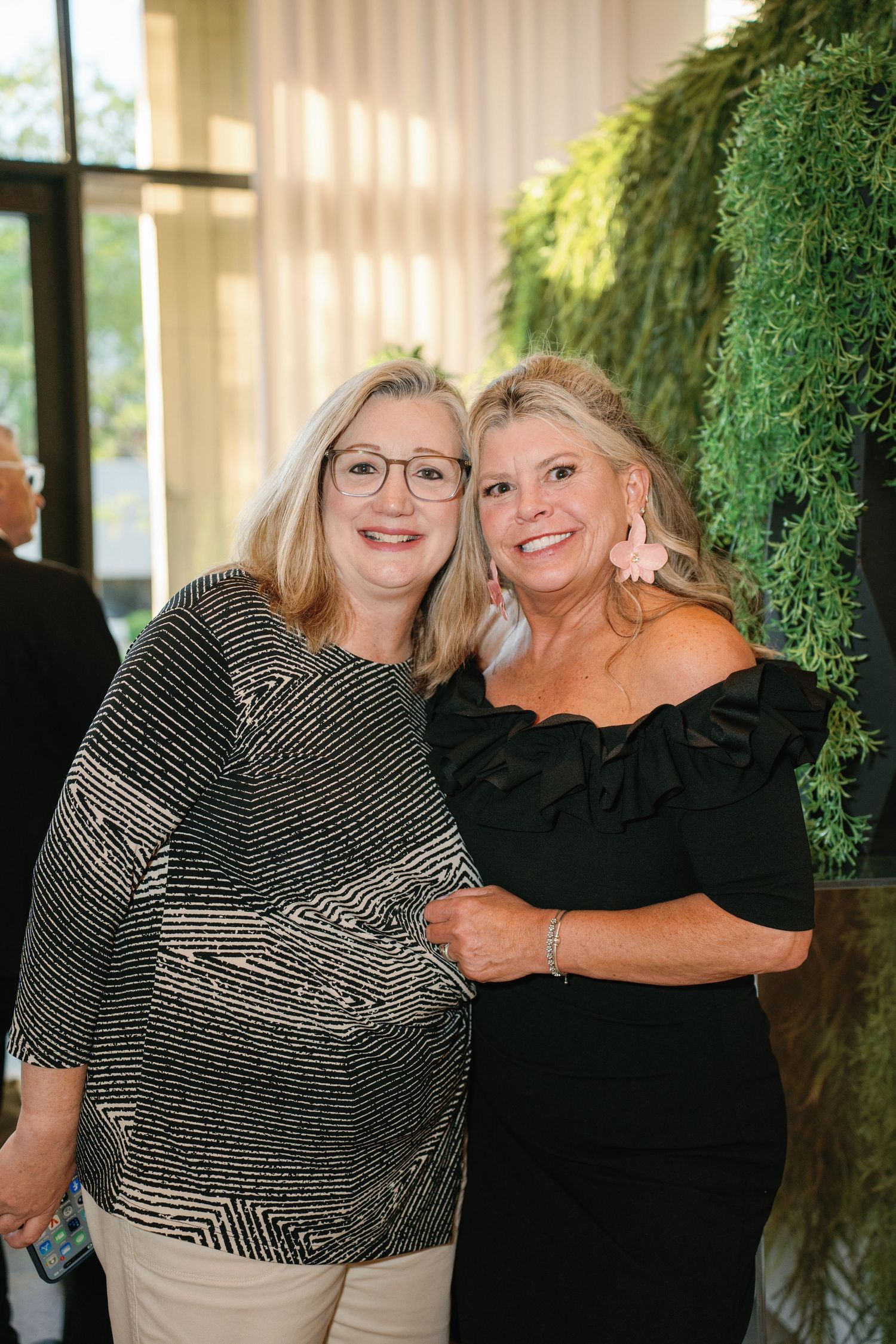 Two smiling women pose together at an elegant event, one in a patterned top and one in a black off-shoulder dress.