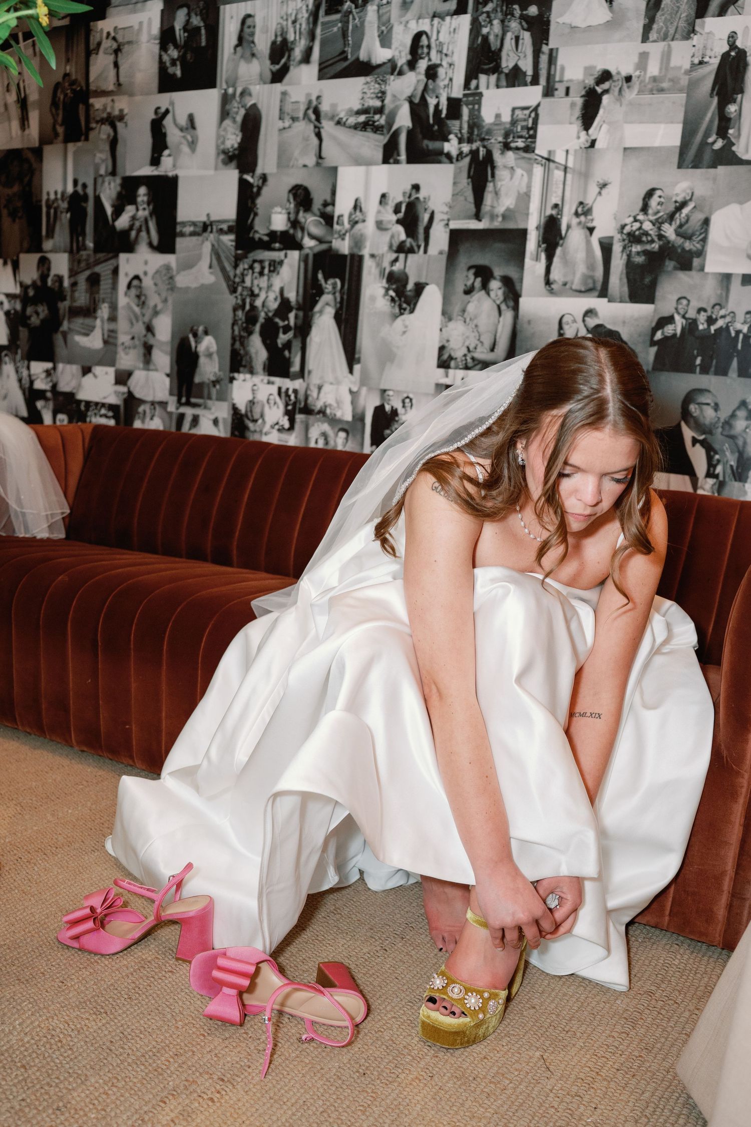 Bride in white gown sits on velvet sofa adjusting gold shoes, pink heels nearby, wedding photos on wall behind her.
