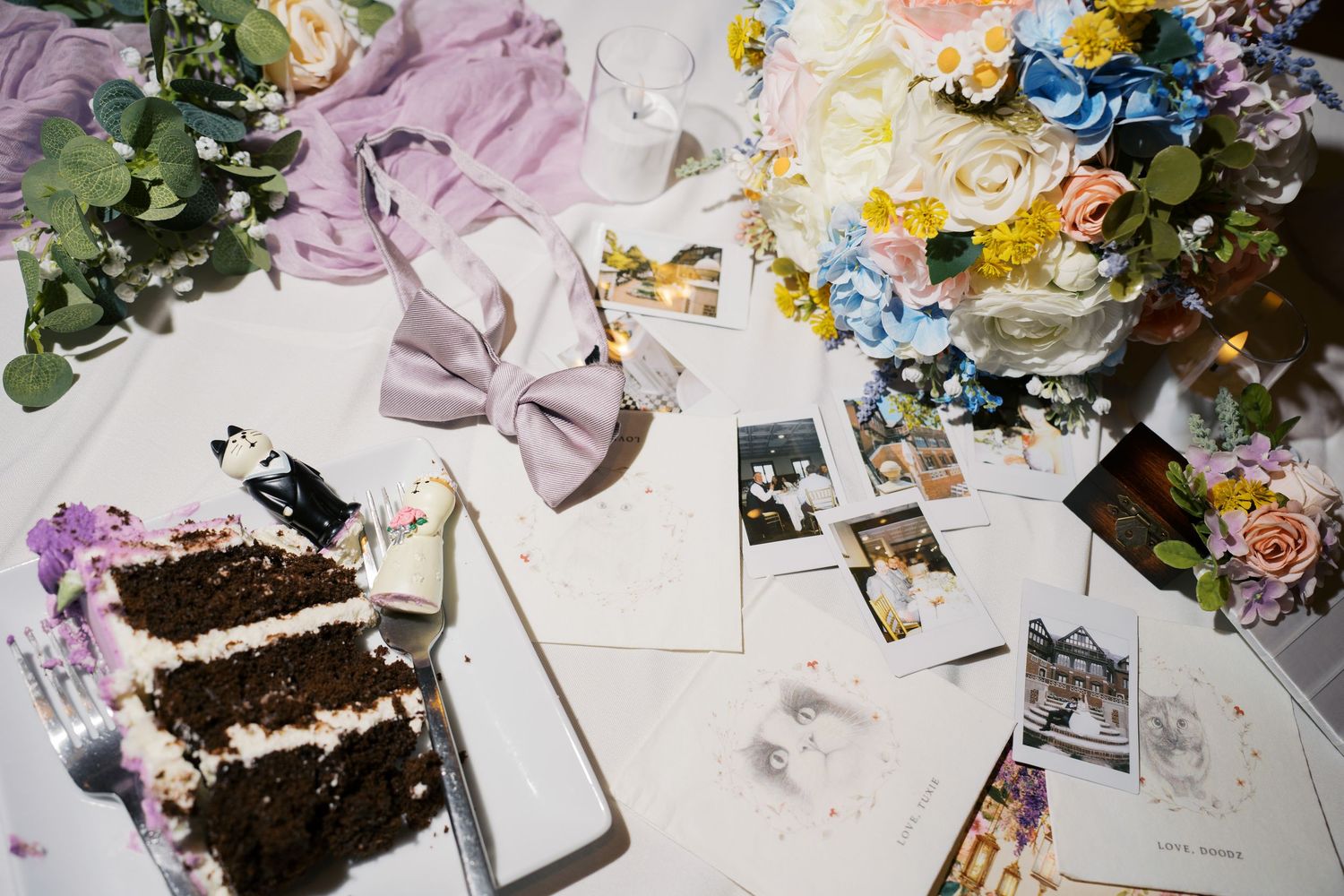 Wedding flat lay with floral bouquets, bow tie, cake slice, Polaroid photos, and stationery on a white table.