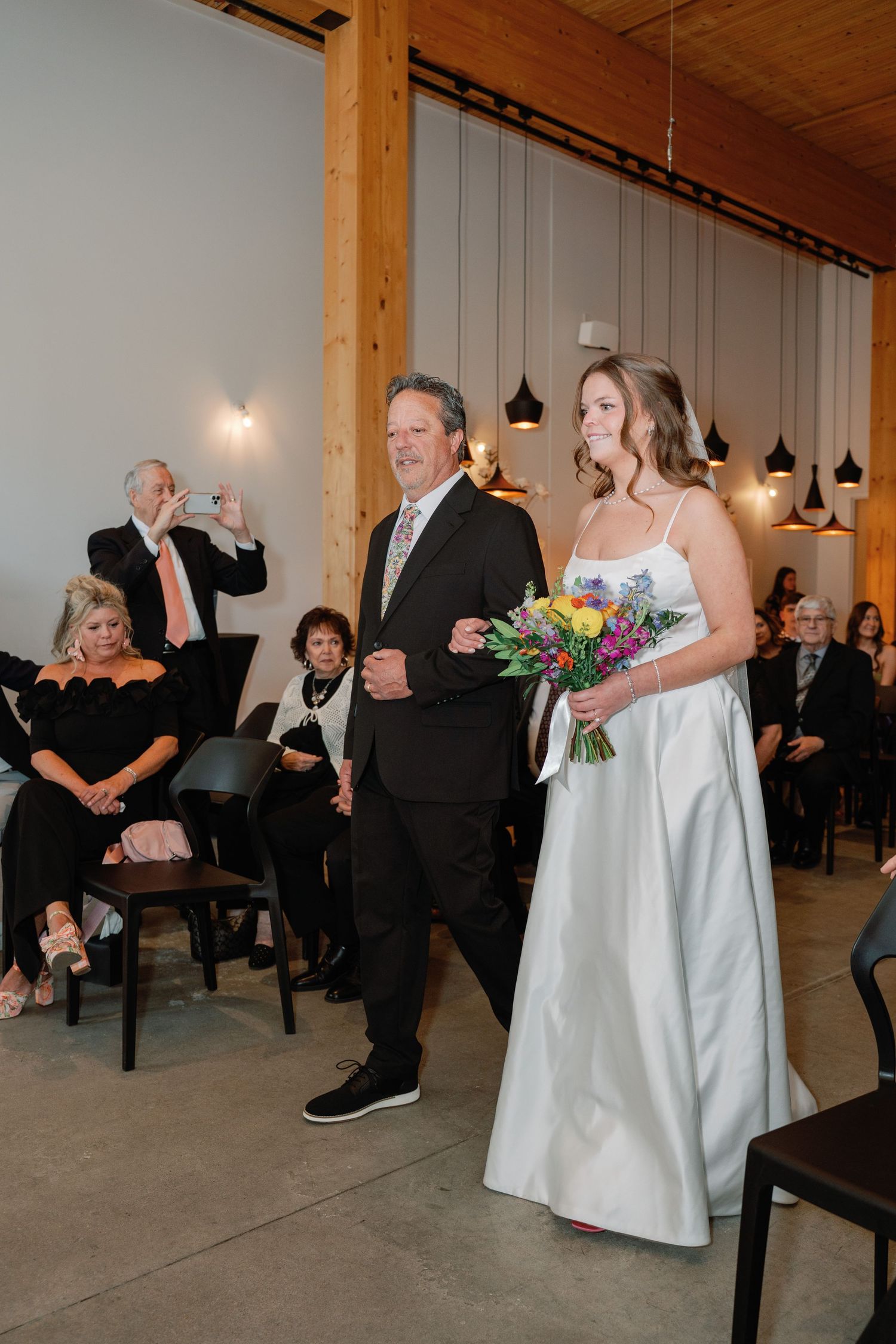 Bride in white gown holding colorful bouquet walks down aisle with escort during indoor wedding ceremony.