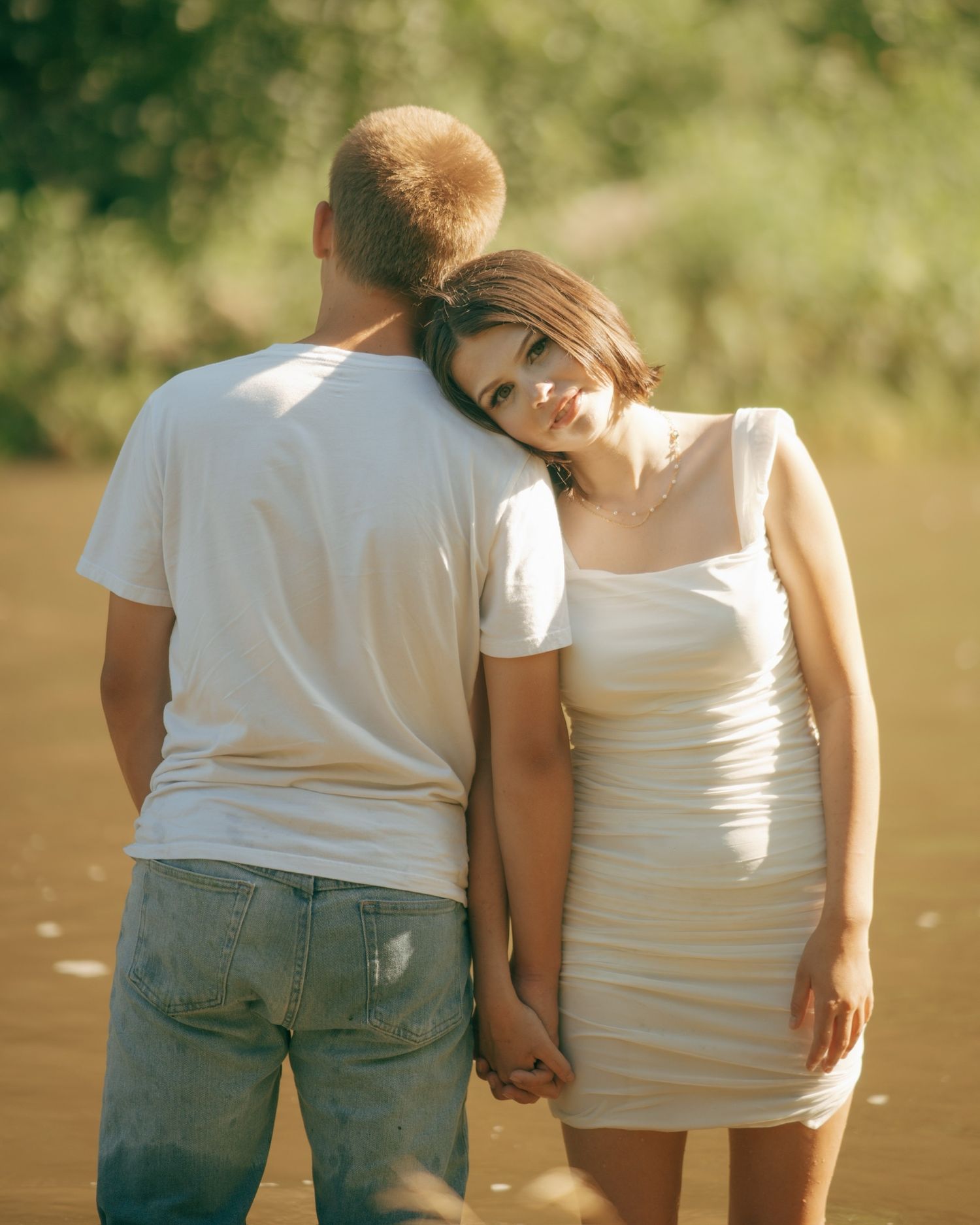 Two people in white shirts and jeans standing together by a lake in warm sunlight.