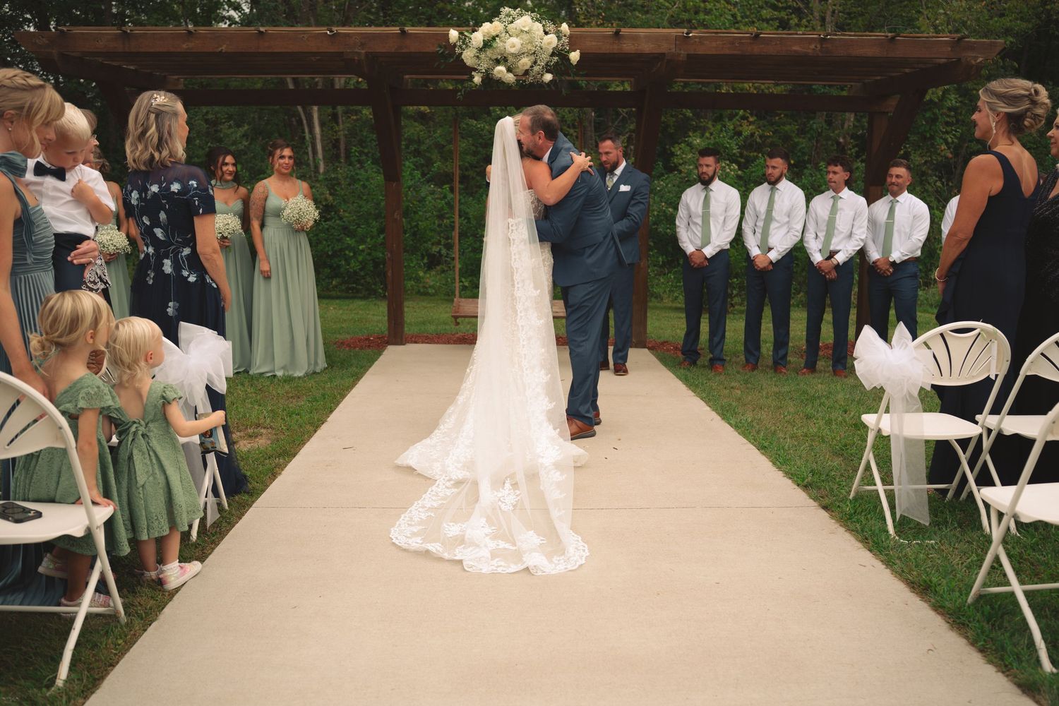 Outdoor wedding ceremony setup with wooden arbor and white chairs under trees.