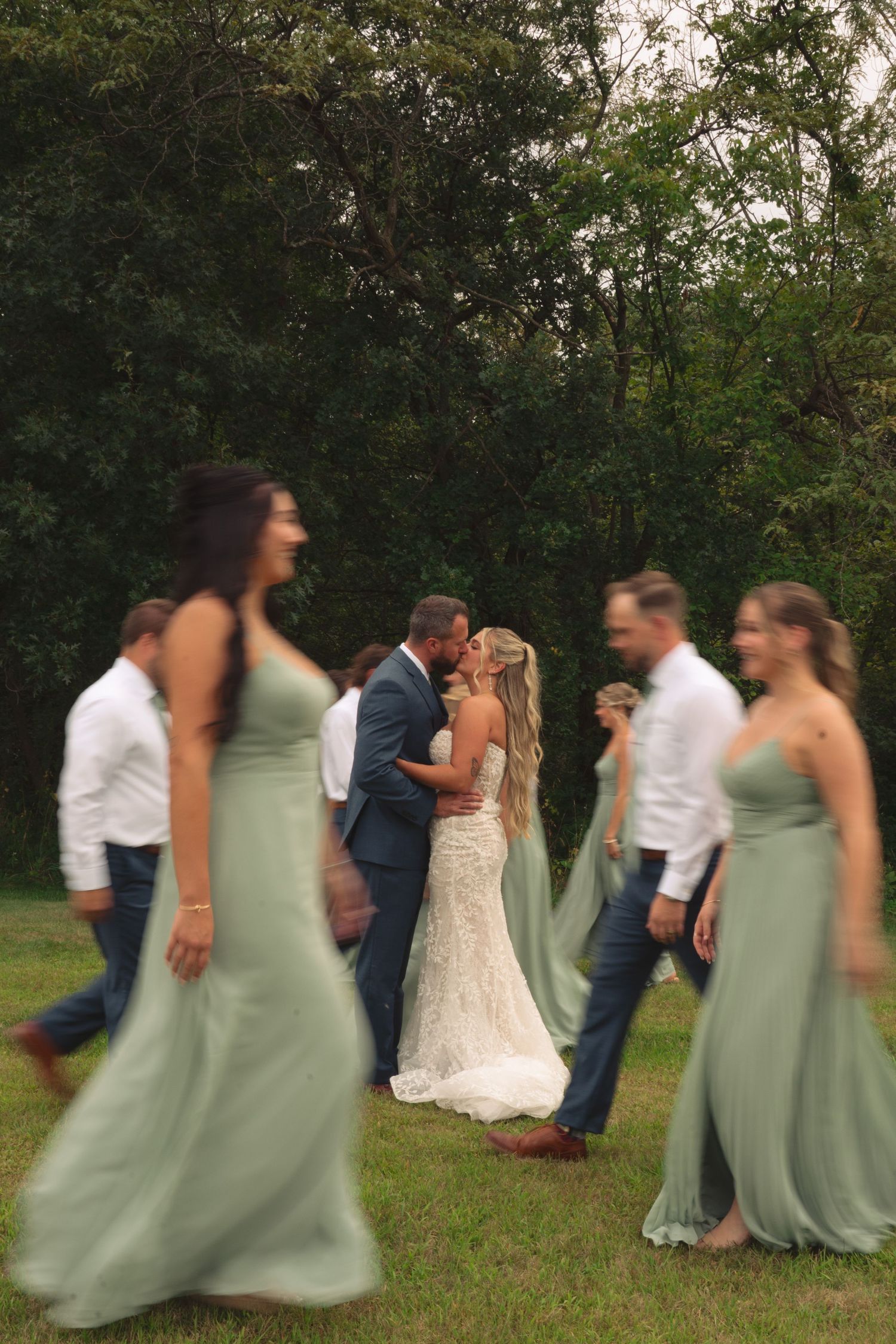 Wedding party in sage green dresses and navy suits poses outdoors for photos.