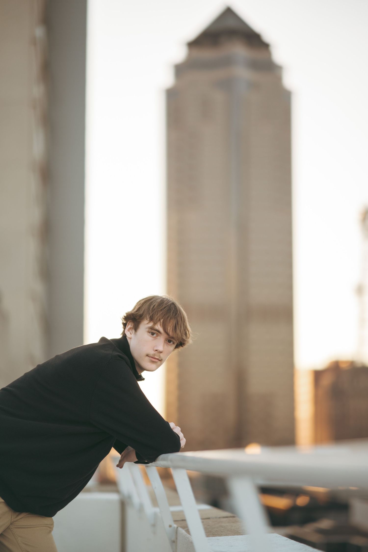 Young man in black jacket leaning on rooftop railing with city skyscraper in background at sunset.