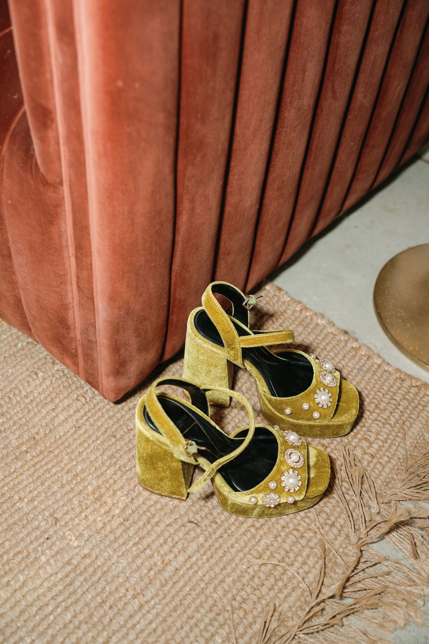 Yellow velvet platform heels with pearl and floral embellishments on woven rug beside pink velvet chair.