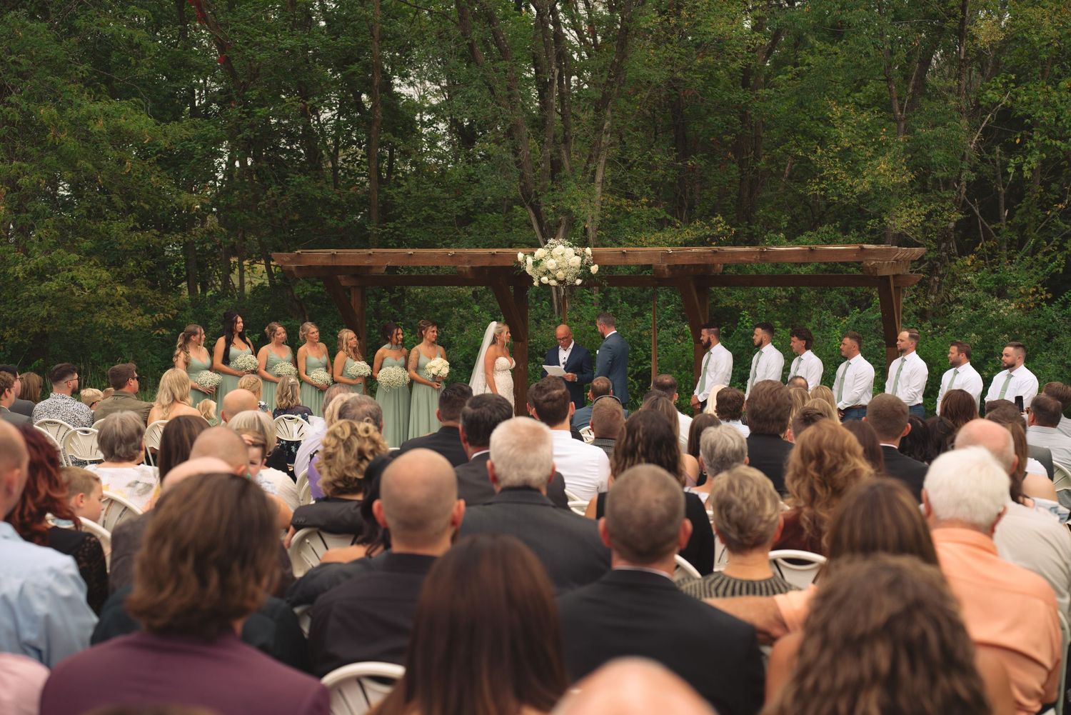 Wedding ceremony in progress under wooden arbor with guests seated on white chairs.