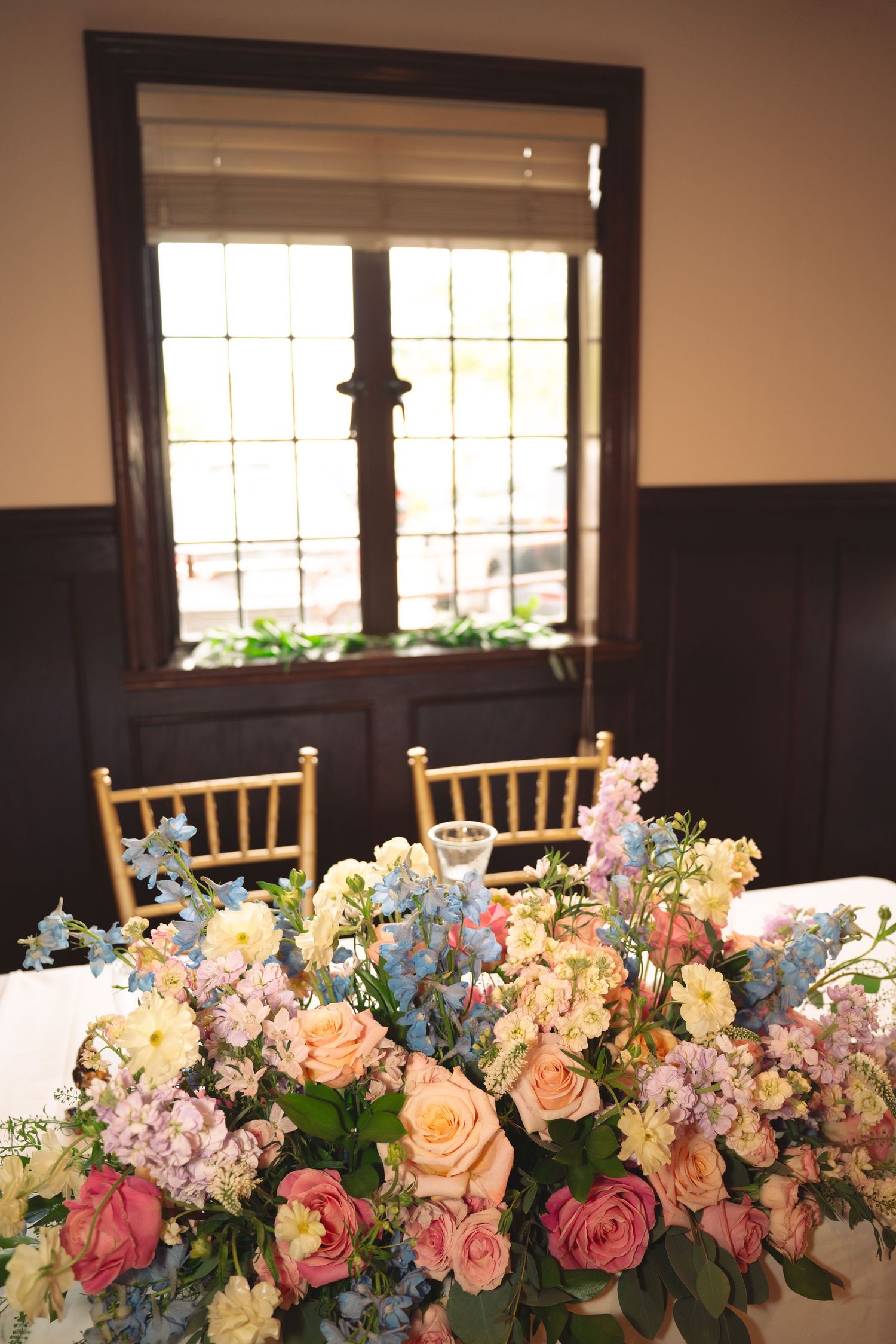 Elegant floral centerpiece with pink, peach and blue flowers against a window in a formal dining setting.