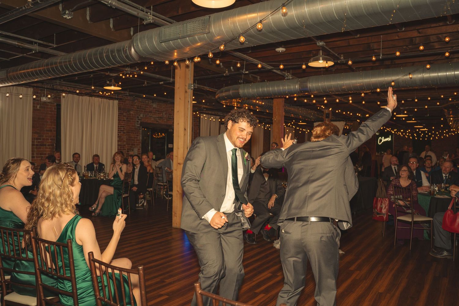Wedding guests dancing on wooden floor under string lights in brick-walled reception venue.
