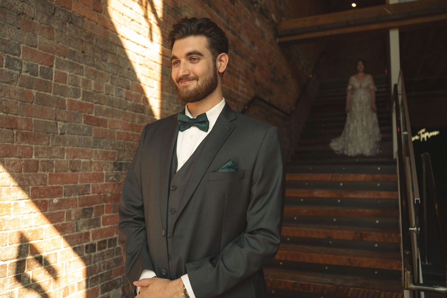 Groom in classic black tuxedo smiles while standing in front of brick wall and staircase.