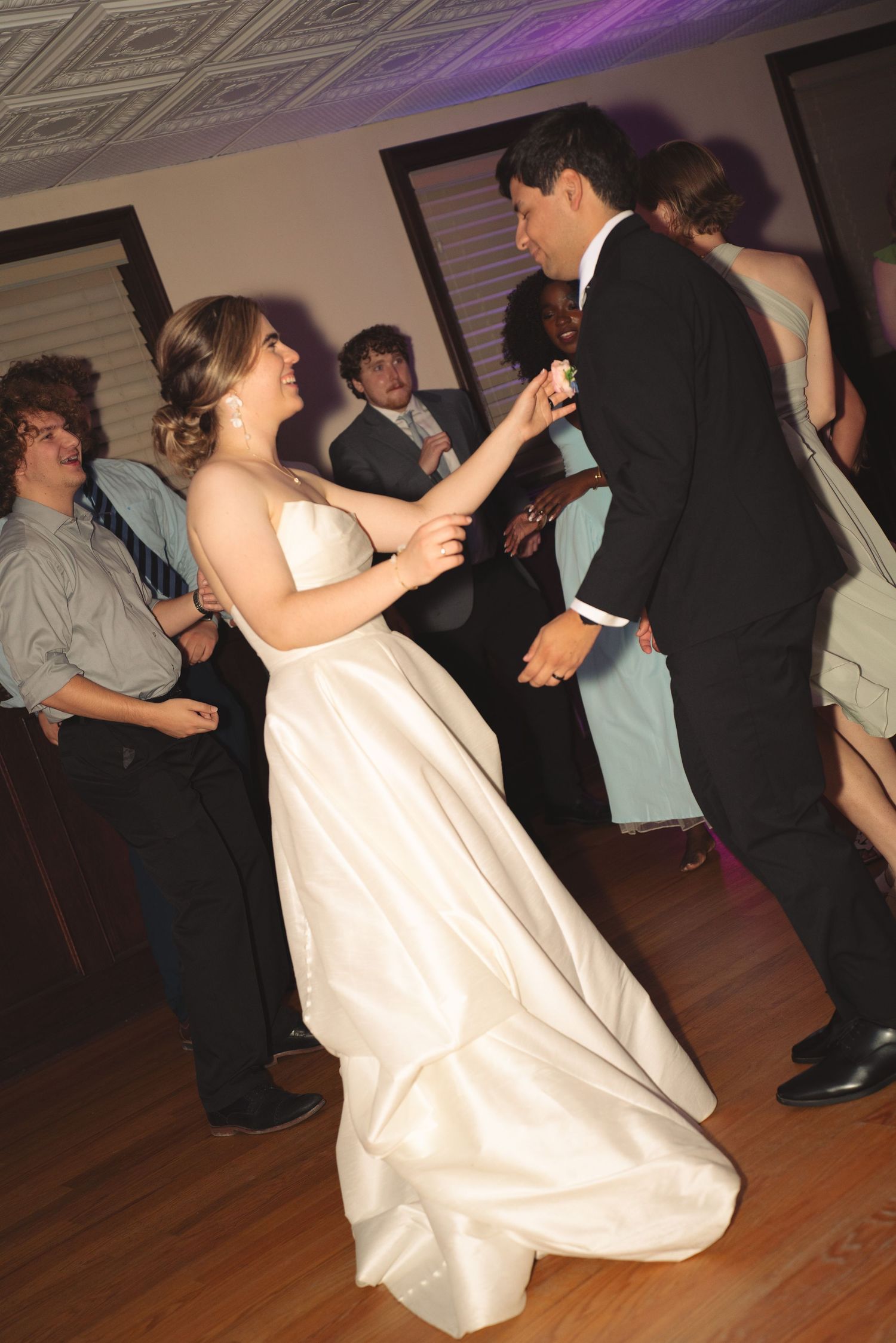 Wedding dancers move across a hardwood floor under purple accent lighting at an indoor venue.