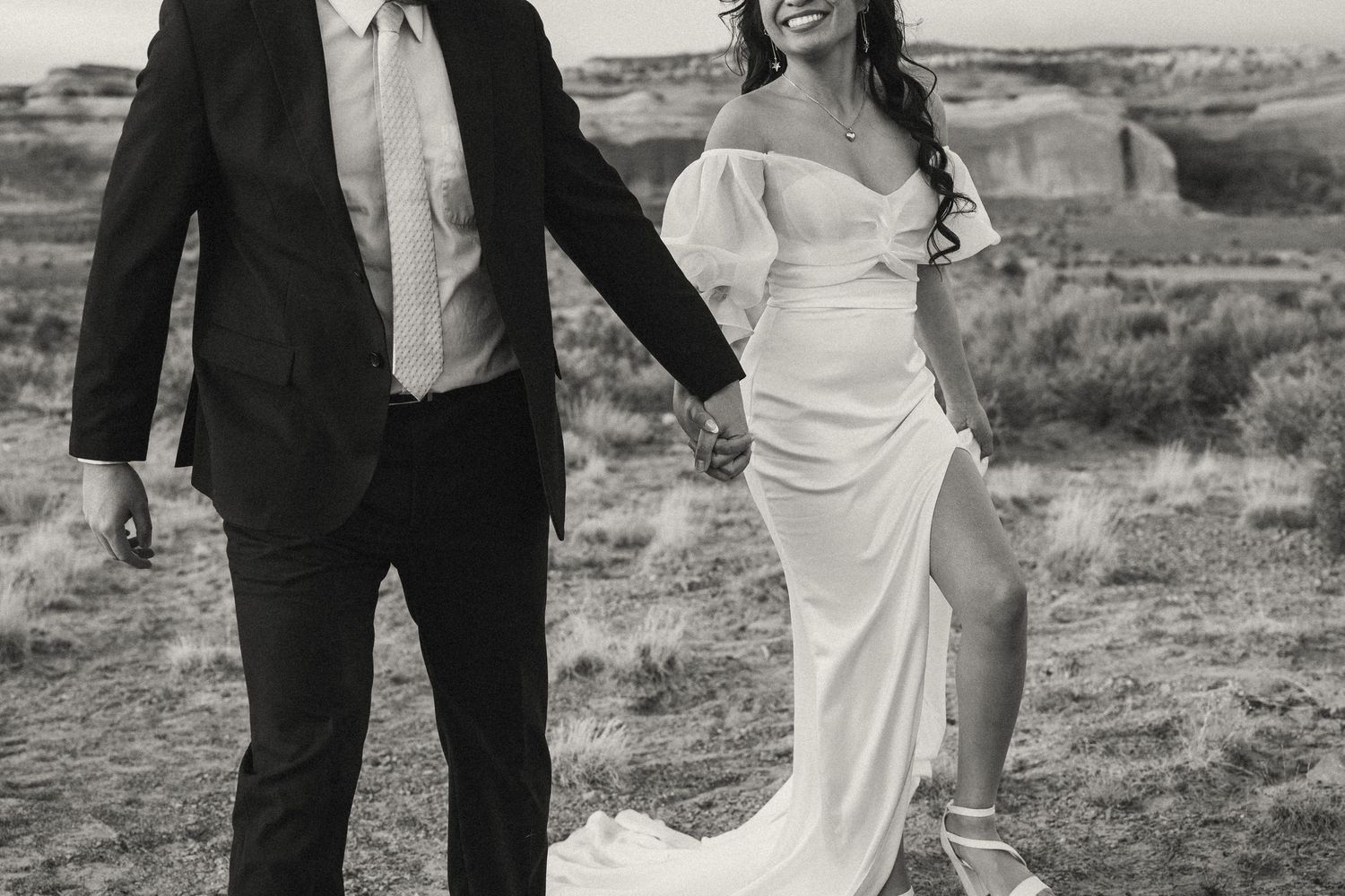 Black and white photo of a smiling bride in white gown and groom in dark suit holding hands outdoors.