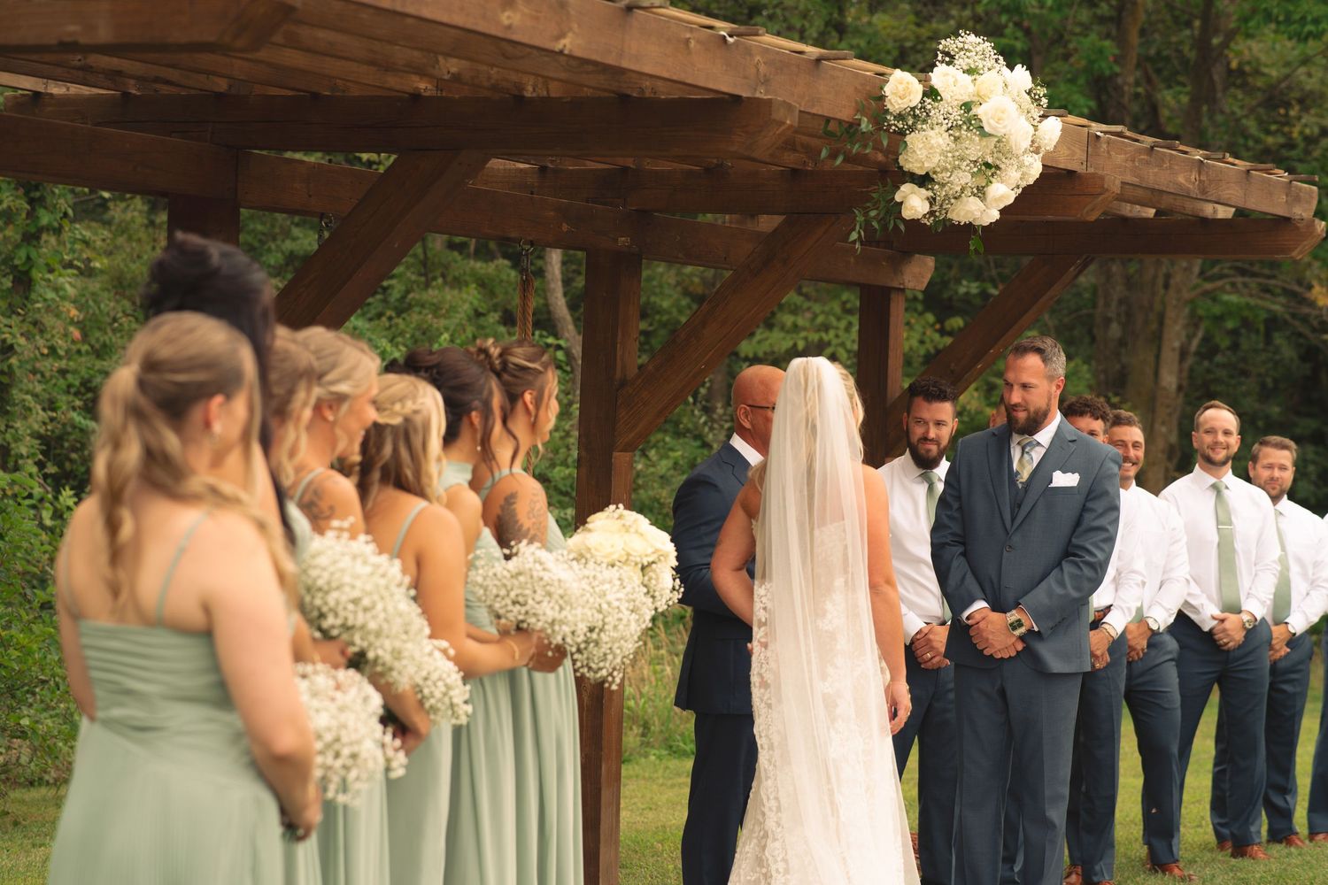 Wedding party stands in formation under a rustic wooden pergola during an outdoor ceremony.