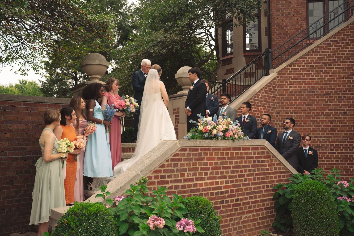 Wedding ceremony guests stand along a brick pathway surrounded by blooming gardens.