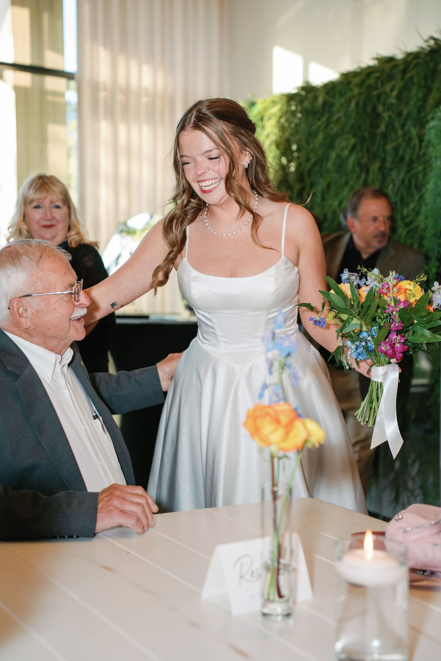 Smiling bride in silver gown holds colorful bouquet while greeting elderly guest at wedding reception table.