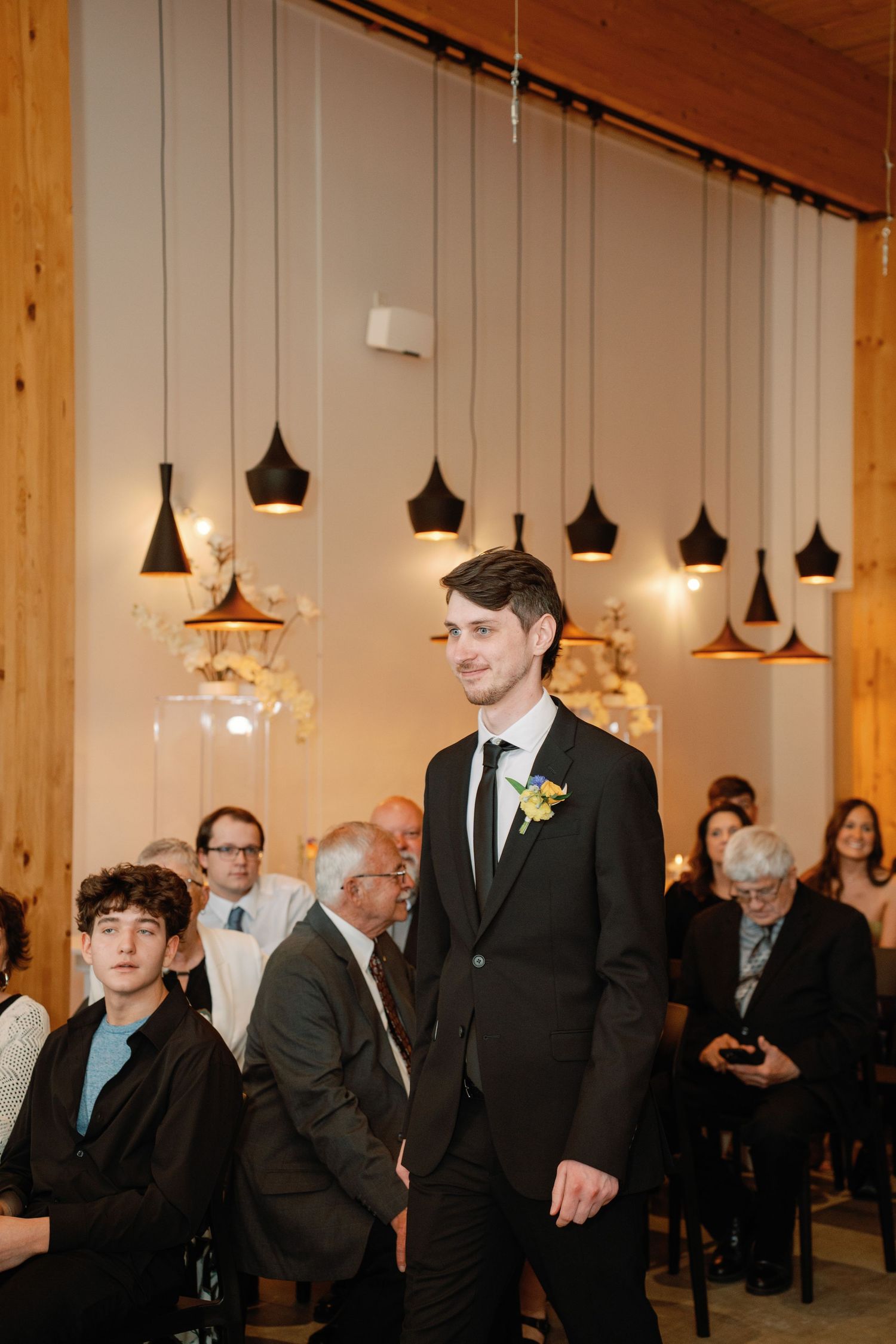 Groom in black suit with yellow boutonniere walks down wedding aisle as guests watch in elegant venue.