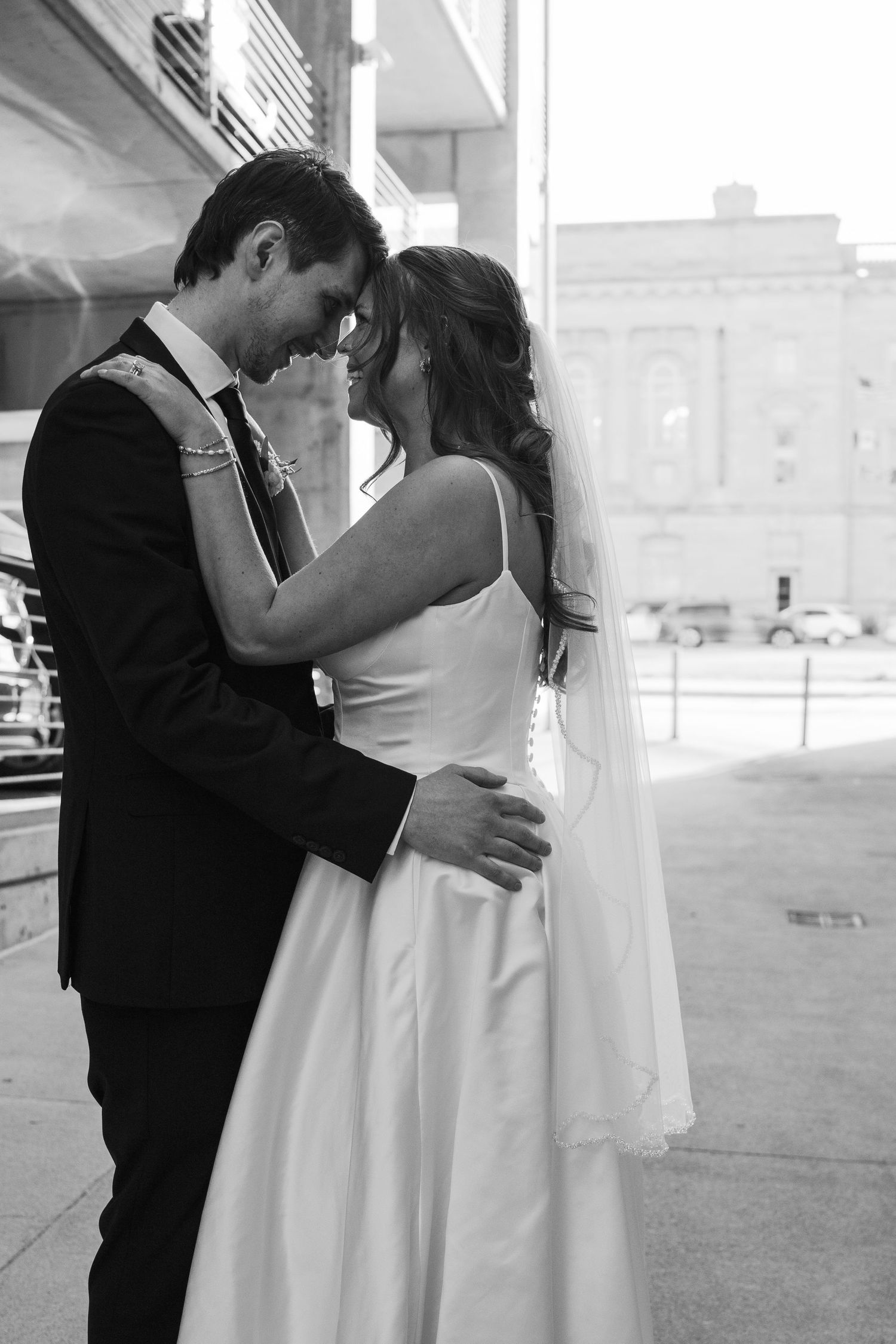 Black and white photo of bride and groom touching foreheads tenderly on urban street in wedding attire.