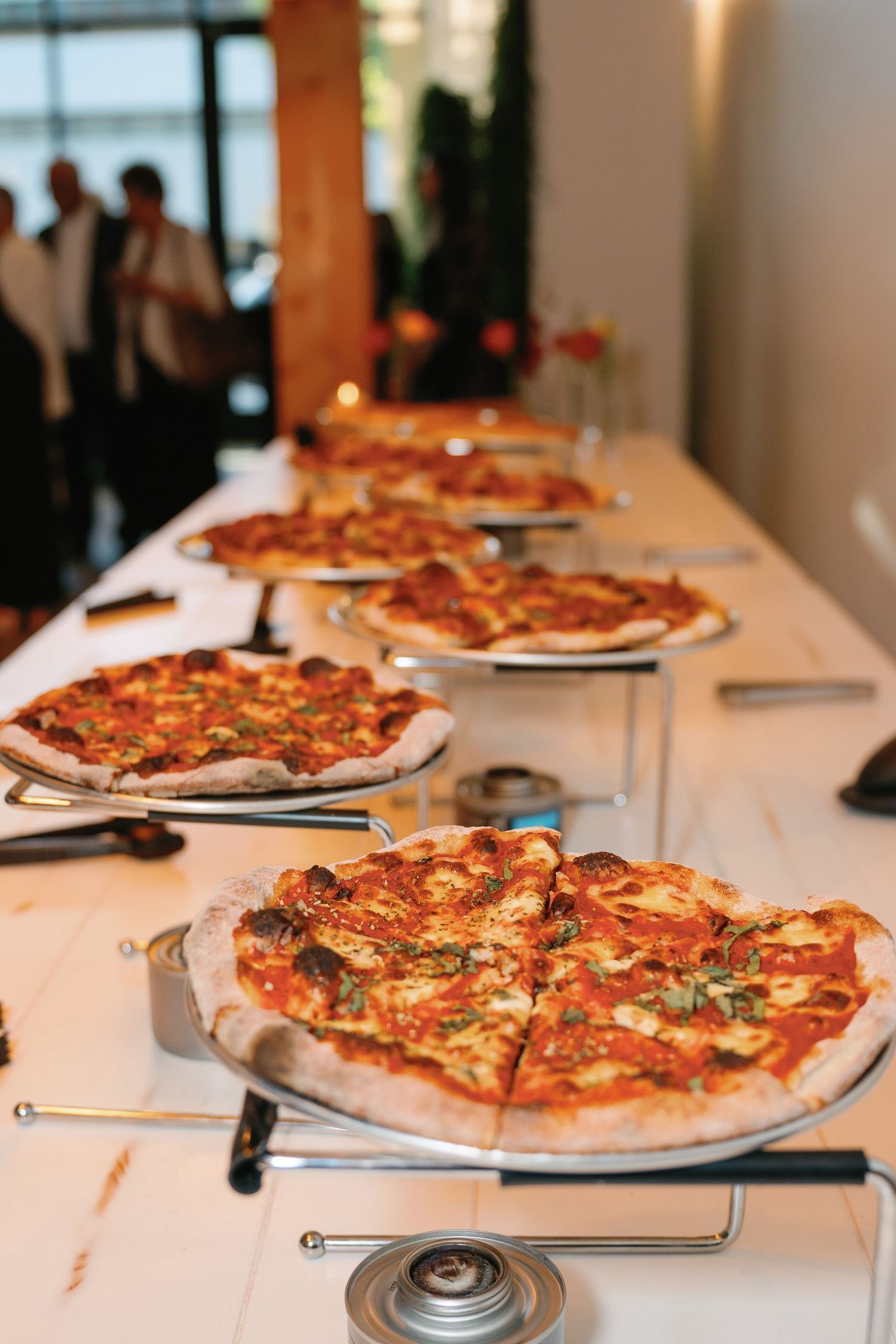 A row of freshly baked pizzas displayed on metal stands along a white table at a catered event.