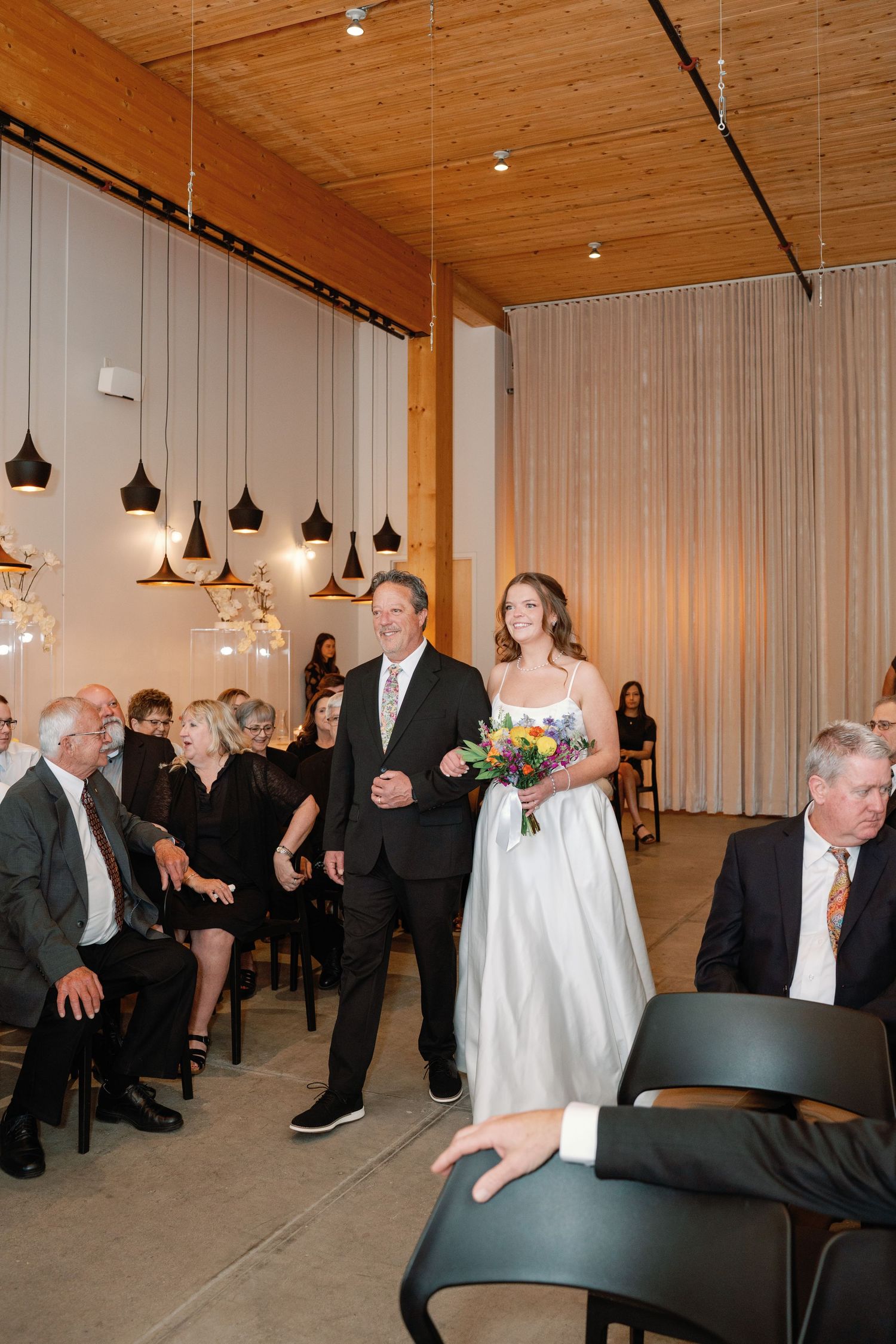 Bride walks down the aisle with her father in a modern venue with pendant lights and seated wedding guests.