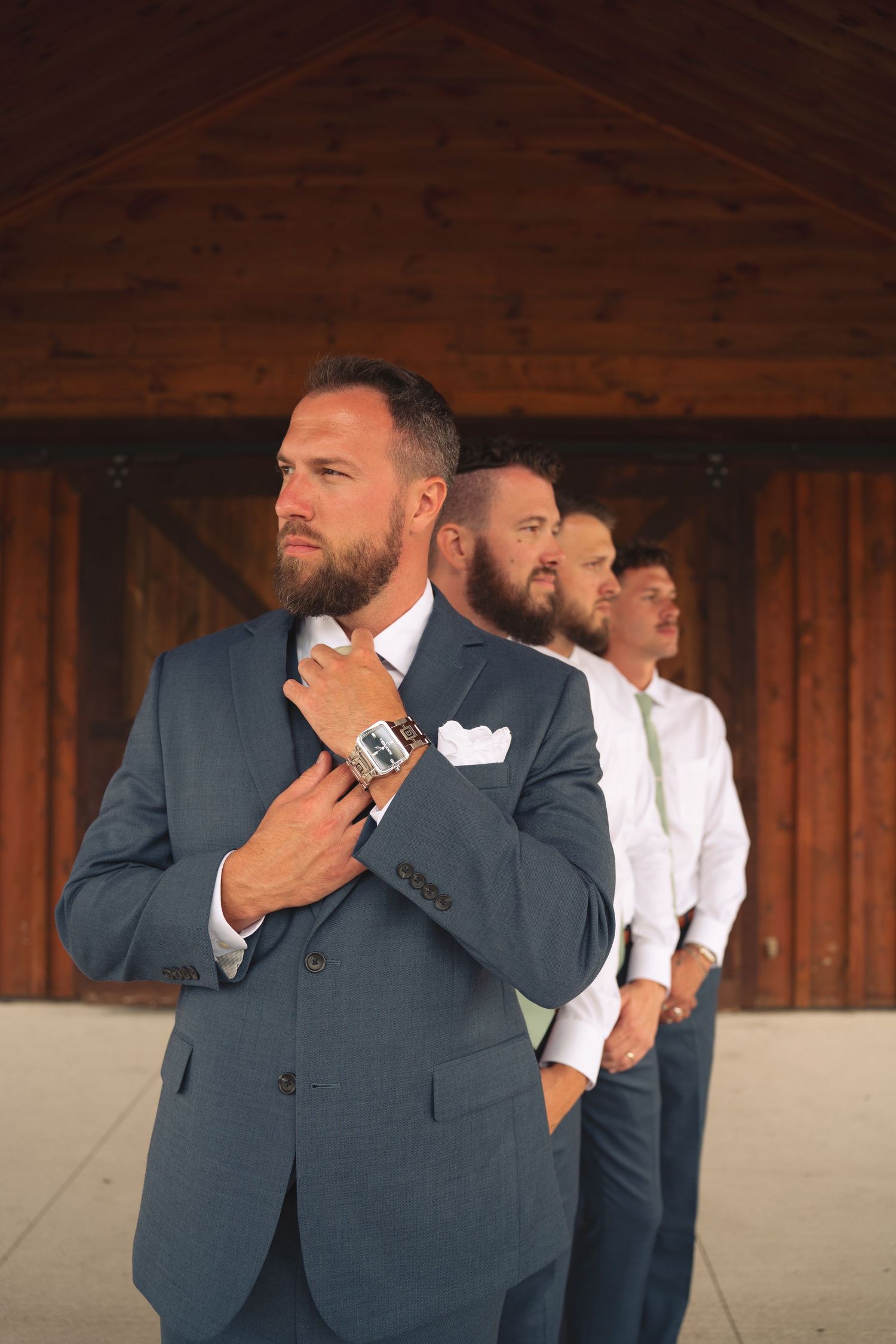 Groomsmen in navy suits adjust their ties while standing in front of wooden barn walls.