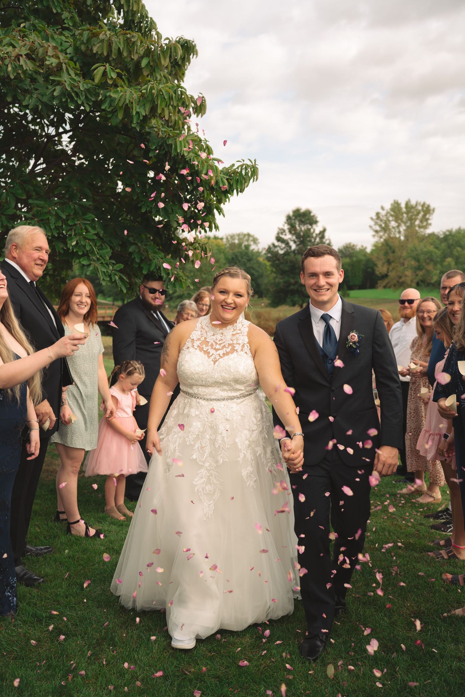 Wedding guests throw rose petals as couple walks through garden celebration.