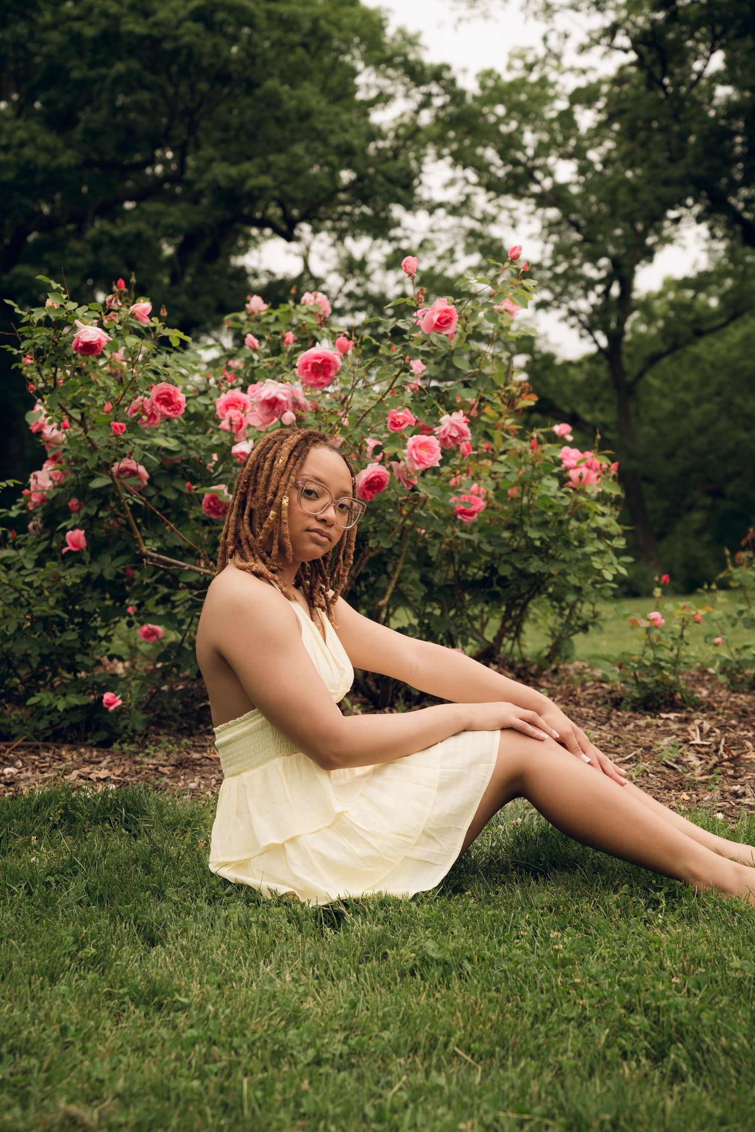 A yellow sundress-clad figure reclines among blooming pink roses in a garden setting.