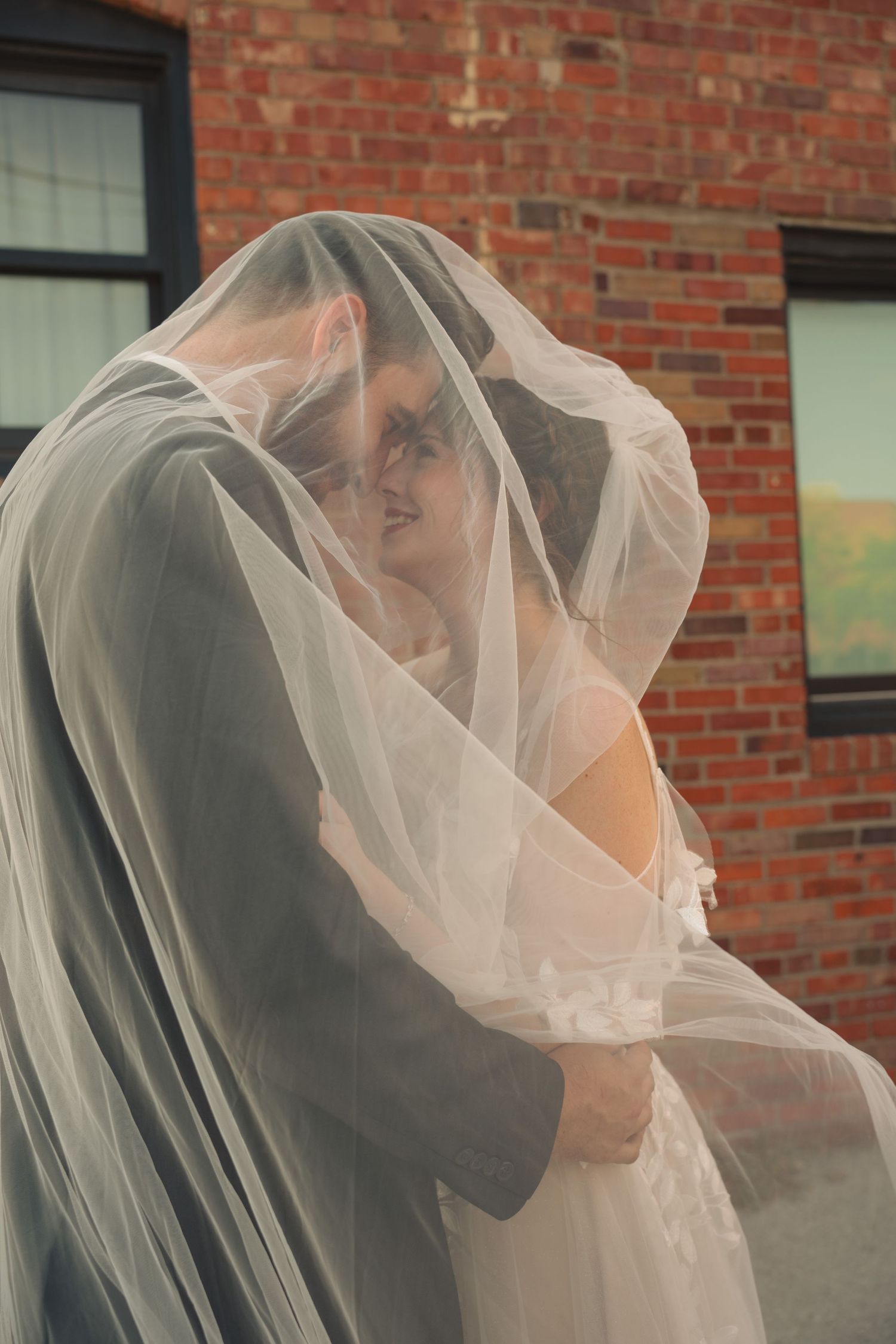 Flowing white wedding veil catches the breeze against a brick building backdrop.
