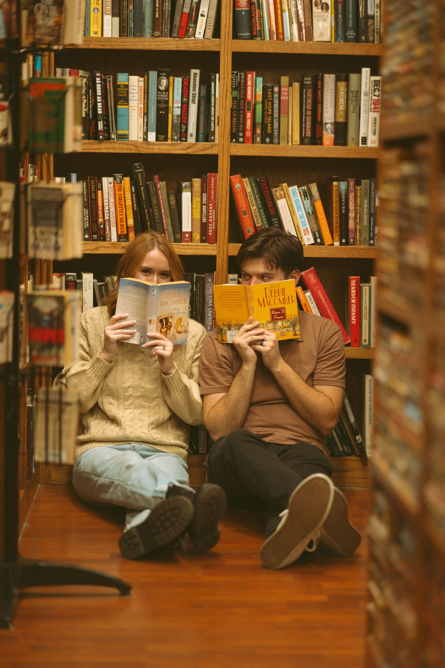 People lying on a wooden floor in a library holding books up to cover their faces.