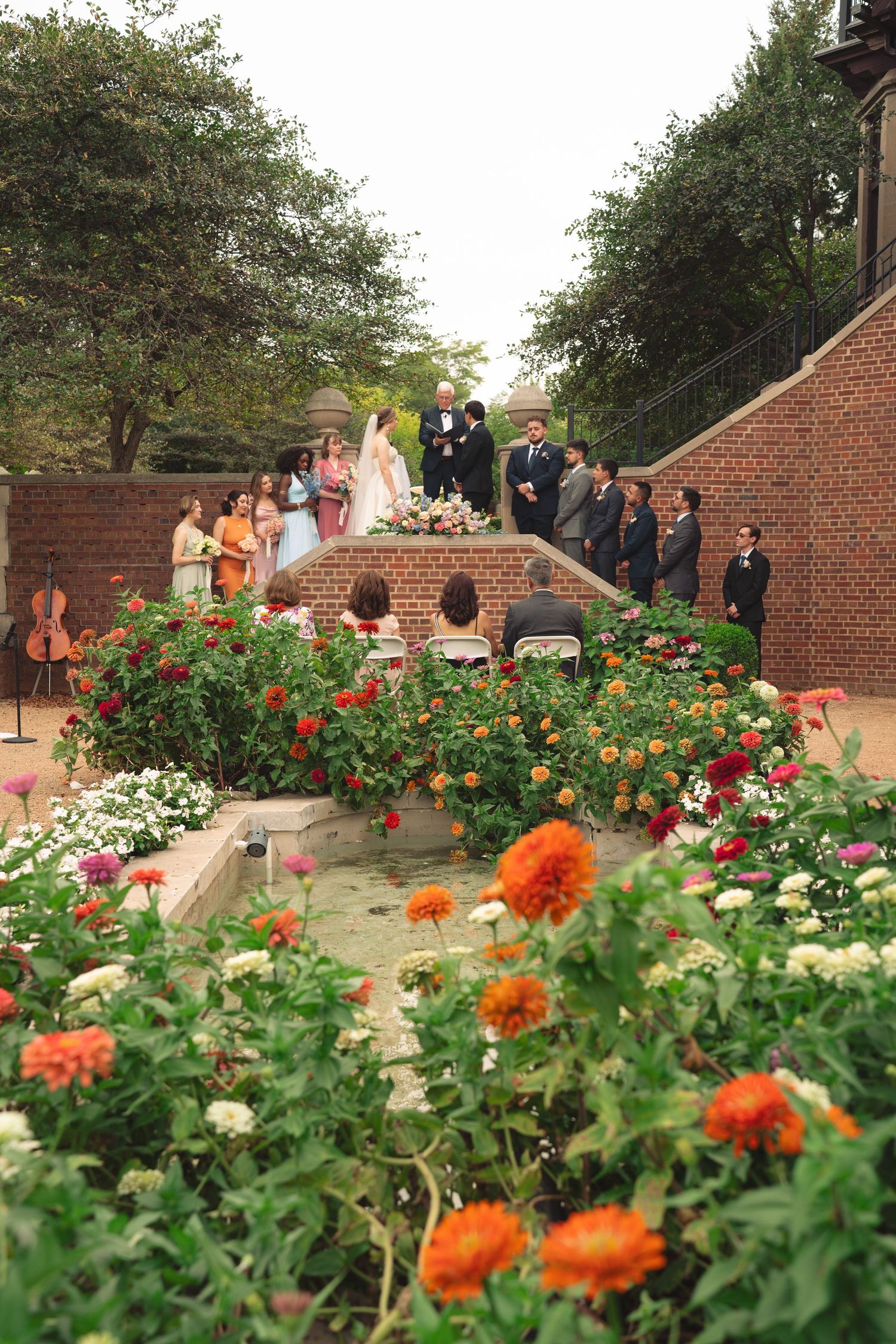 Colorful zinnias and marigolds bloom in a garden bed with wedding guests visible in background.