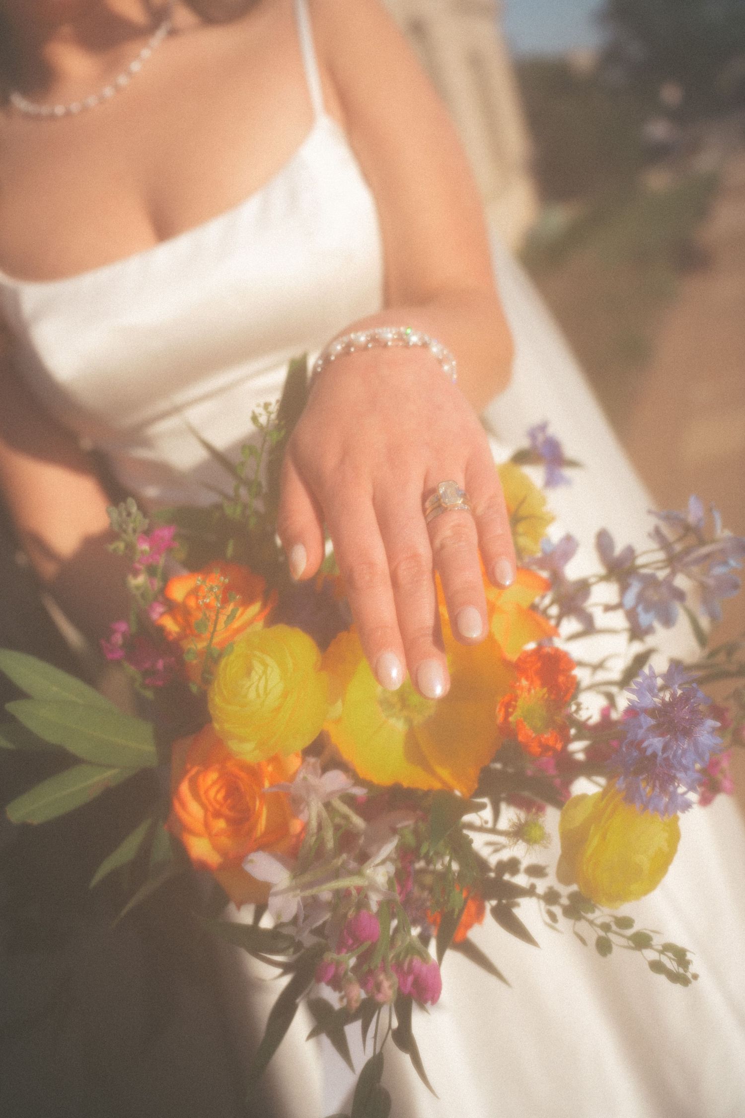 Bride displaying wedding ring and bracelet over colorful bridal bouquet with orange and yellow flowers in white dress.