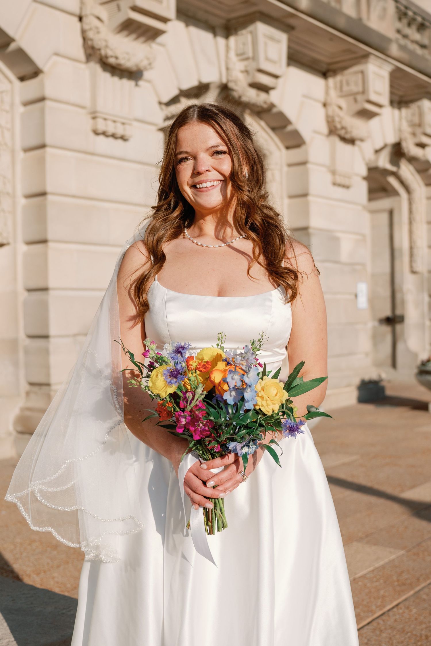 Smiling bride in white strapless gown holding colorful bouquet of yellow, purple, and blue flowers outside elegant stone building.