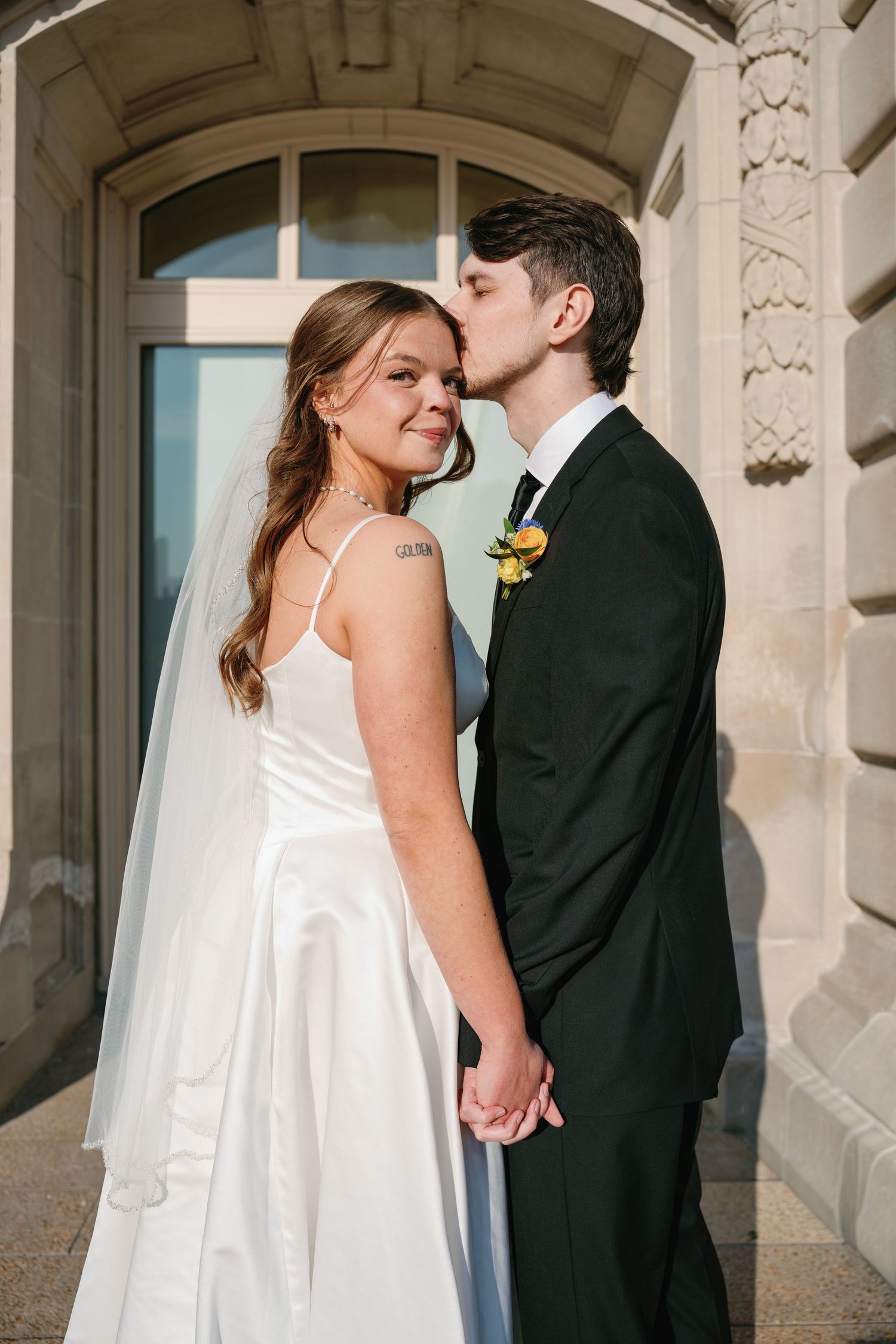 Groom in black suit kisses bride's temple as she smiles, holding hands outside ornate stone building.