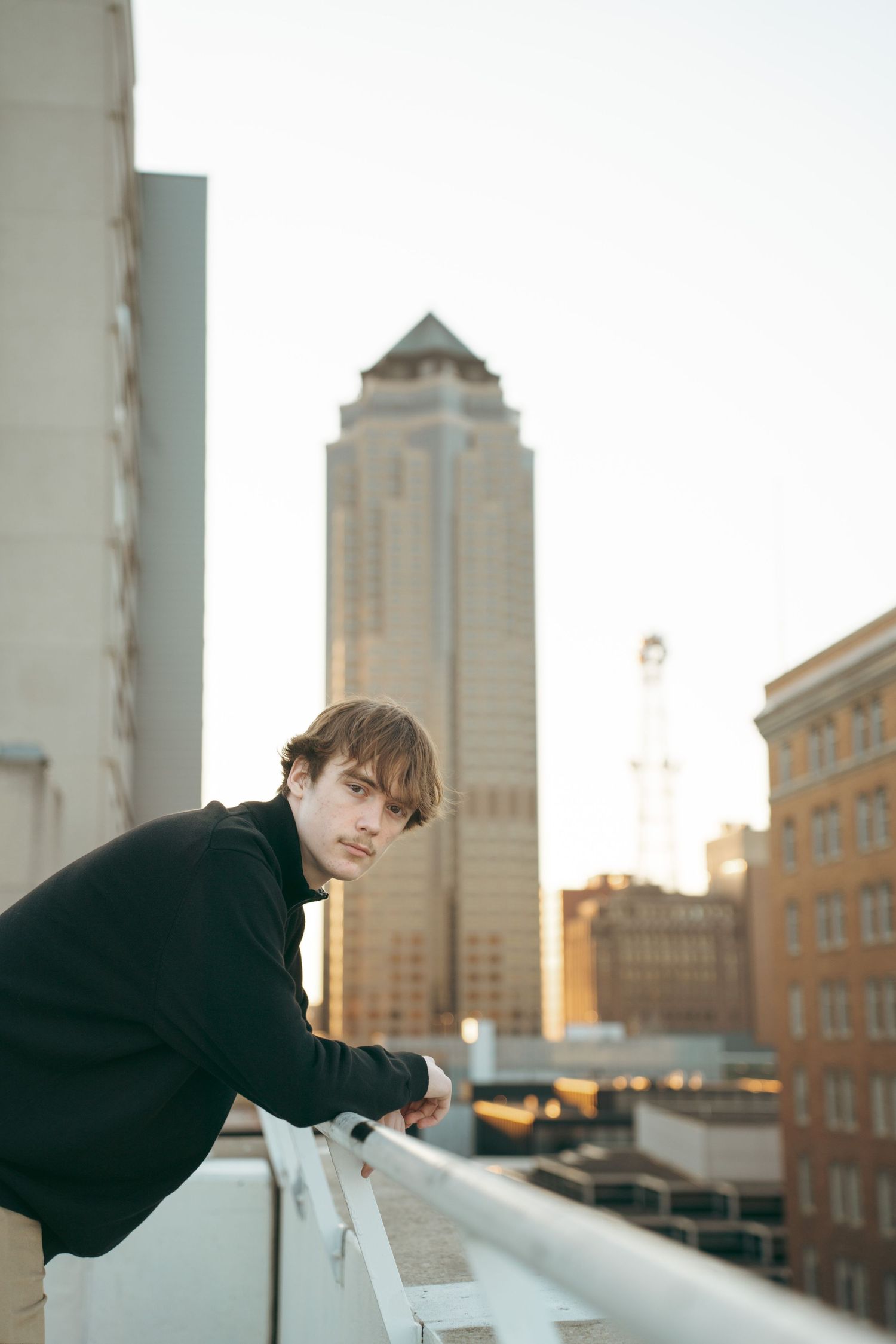 Person in black coat leans against railing on urban rooftop with tall skyscraper visible in background at sunset.