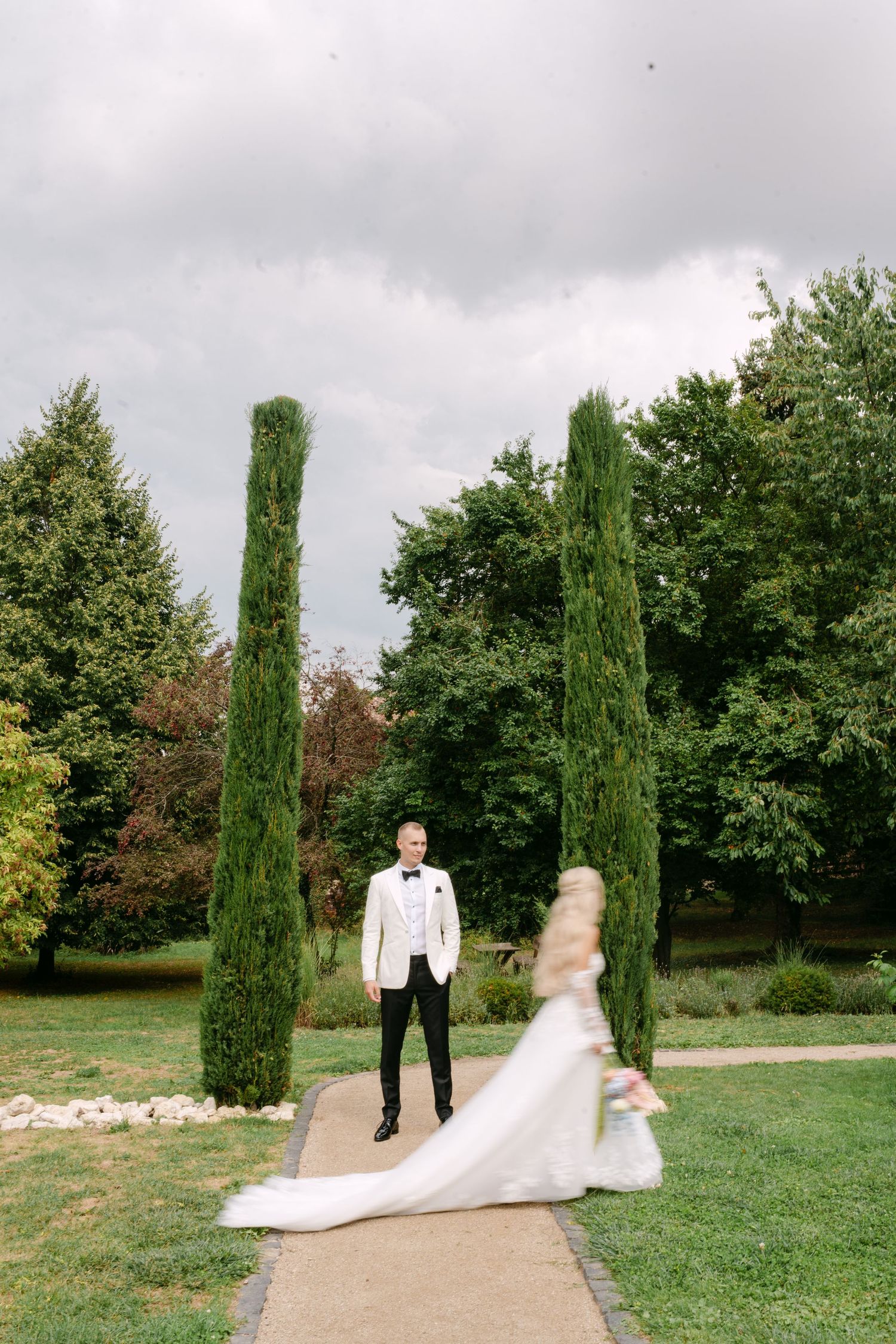 A couple walks along a garden path lined with tall cypress trees on their wedding day.