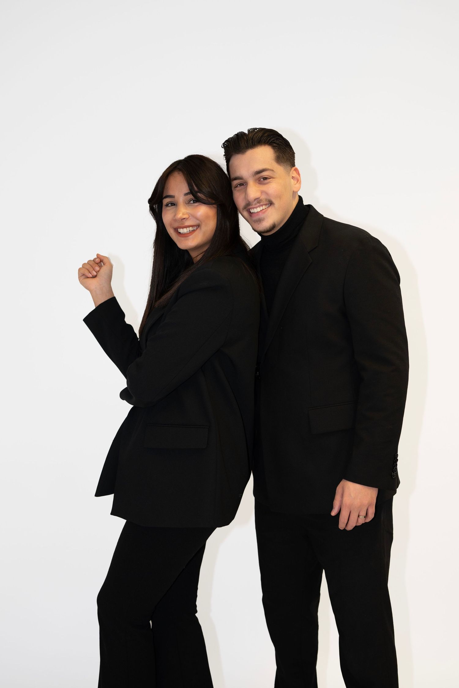 Two people in black clothing smile and pose playfully against a white studio background.