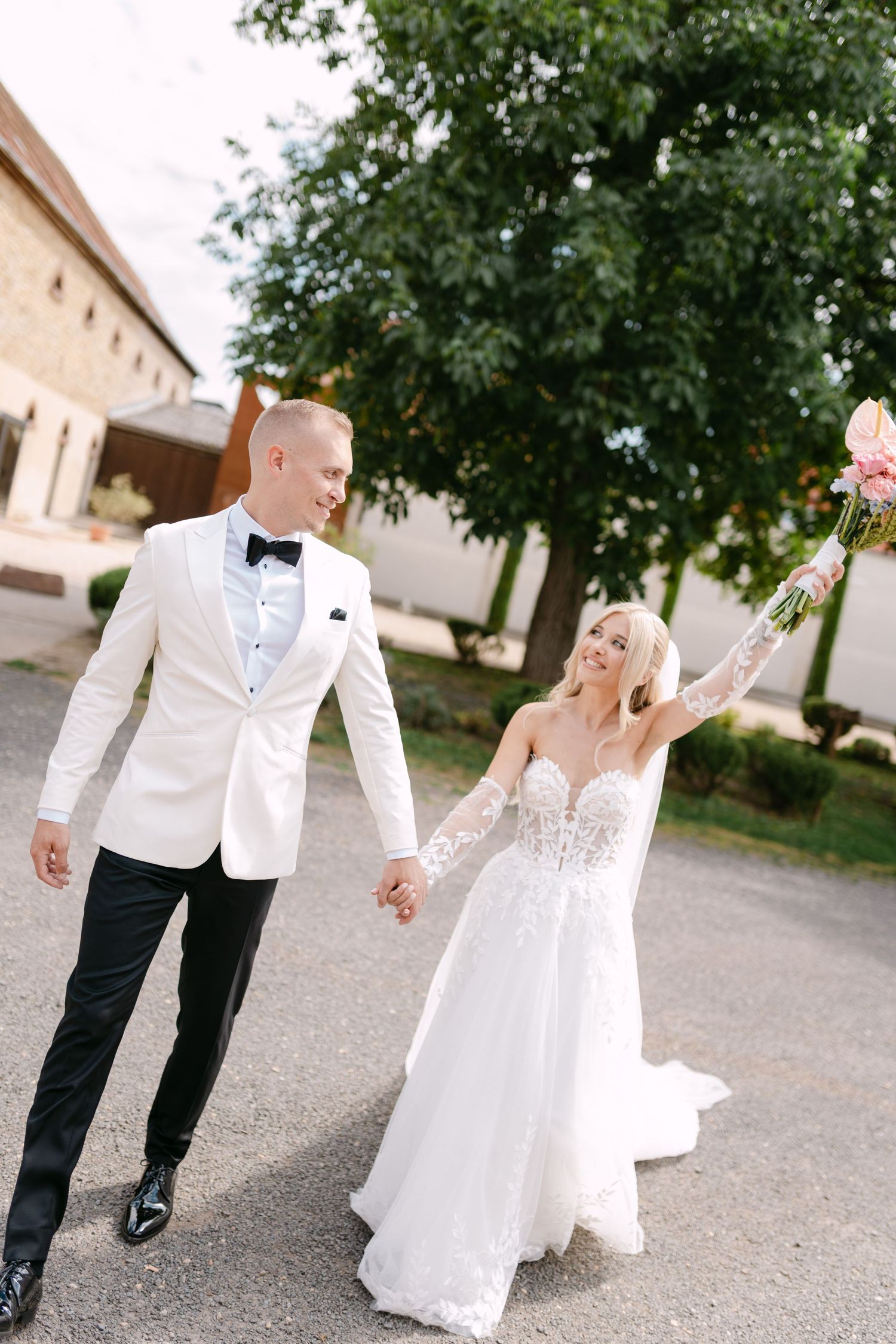 Newlyweds walking hand in hand on a stone pathway surrounded by green trees.