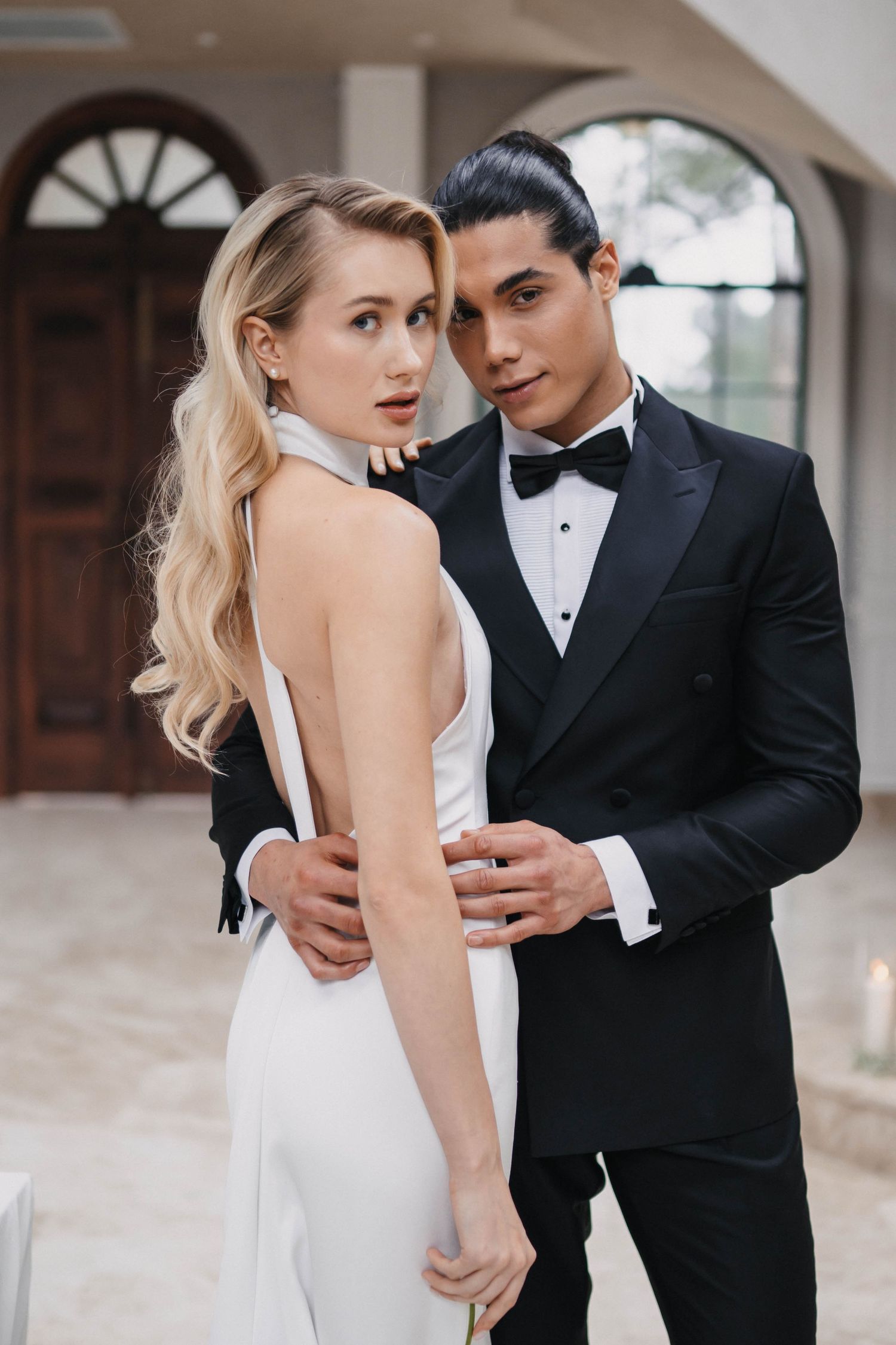 Candid wedding portraits of a couple in black and white formal attire embracing near arched windows.