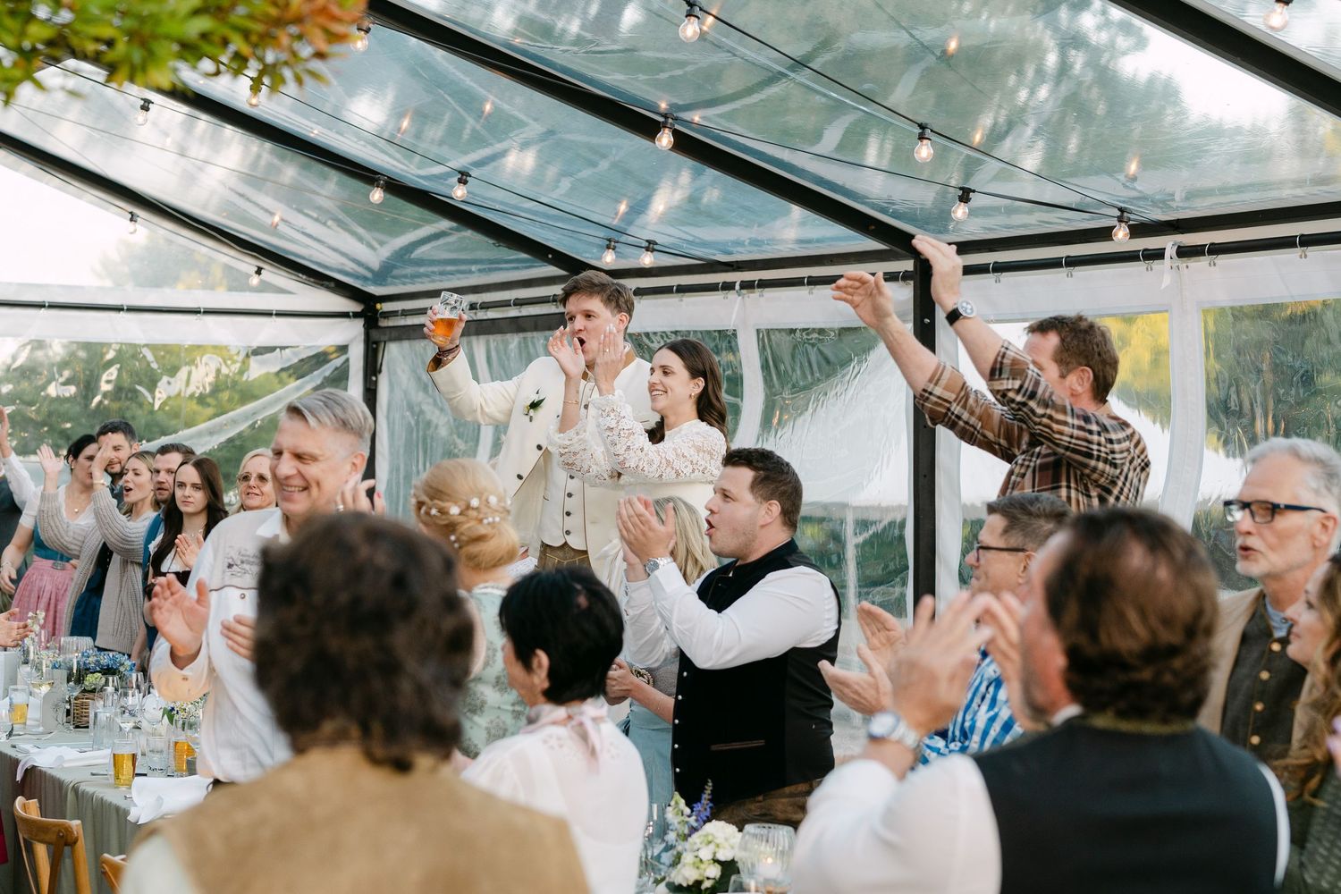 Wedding guests dancing and celebrating under a clear tent with string lights.