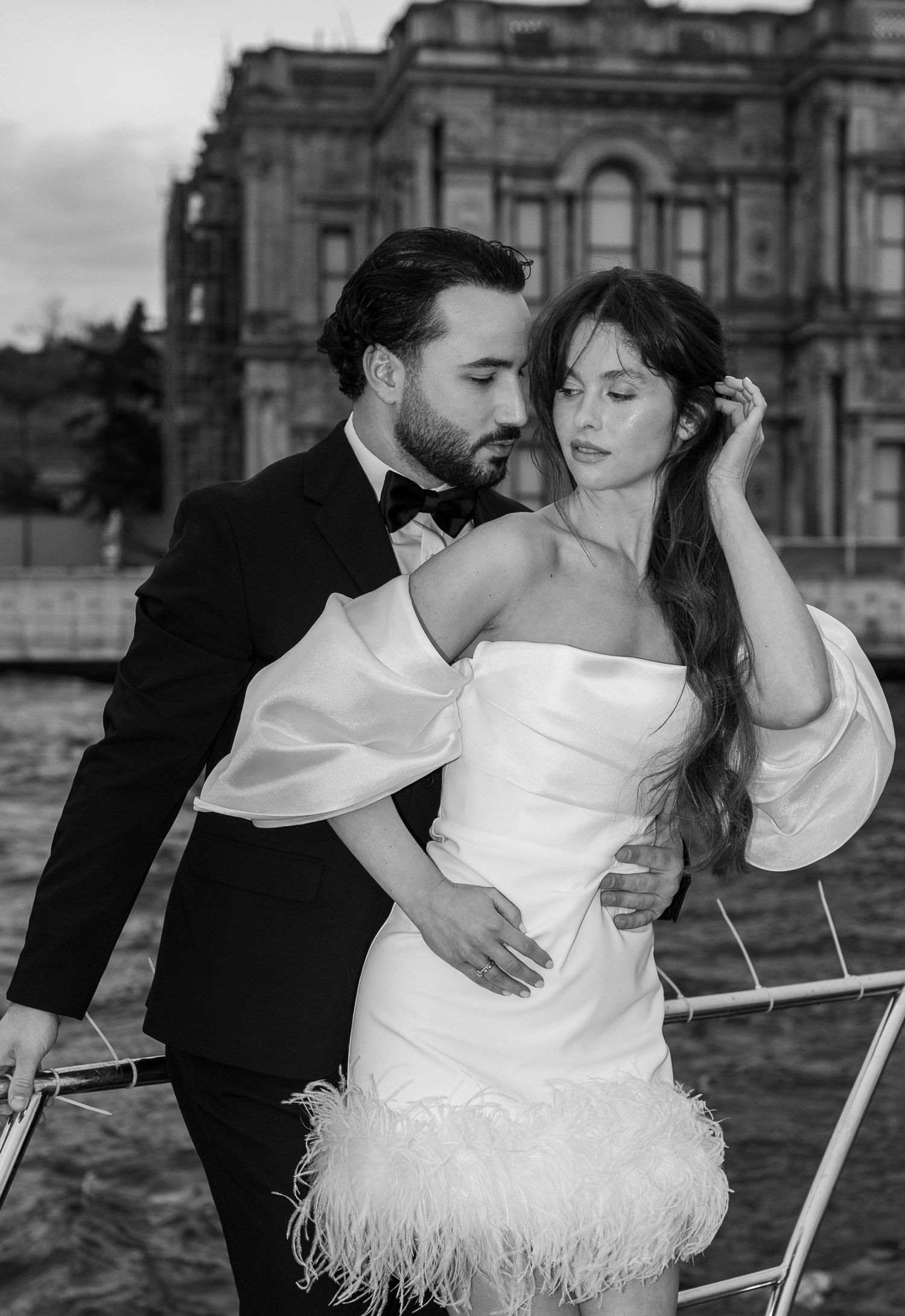 Romantic couple embracing on boat with historic building backdrop in black and white photography.