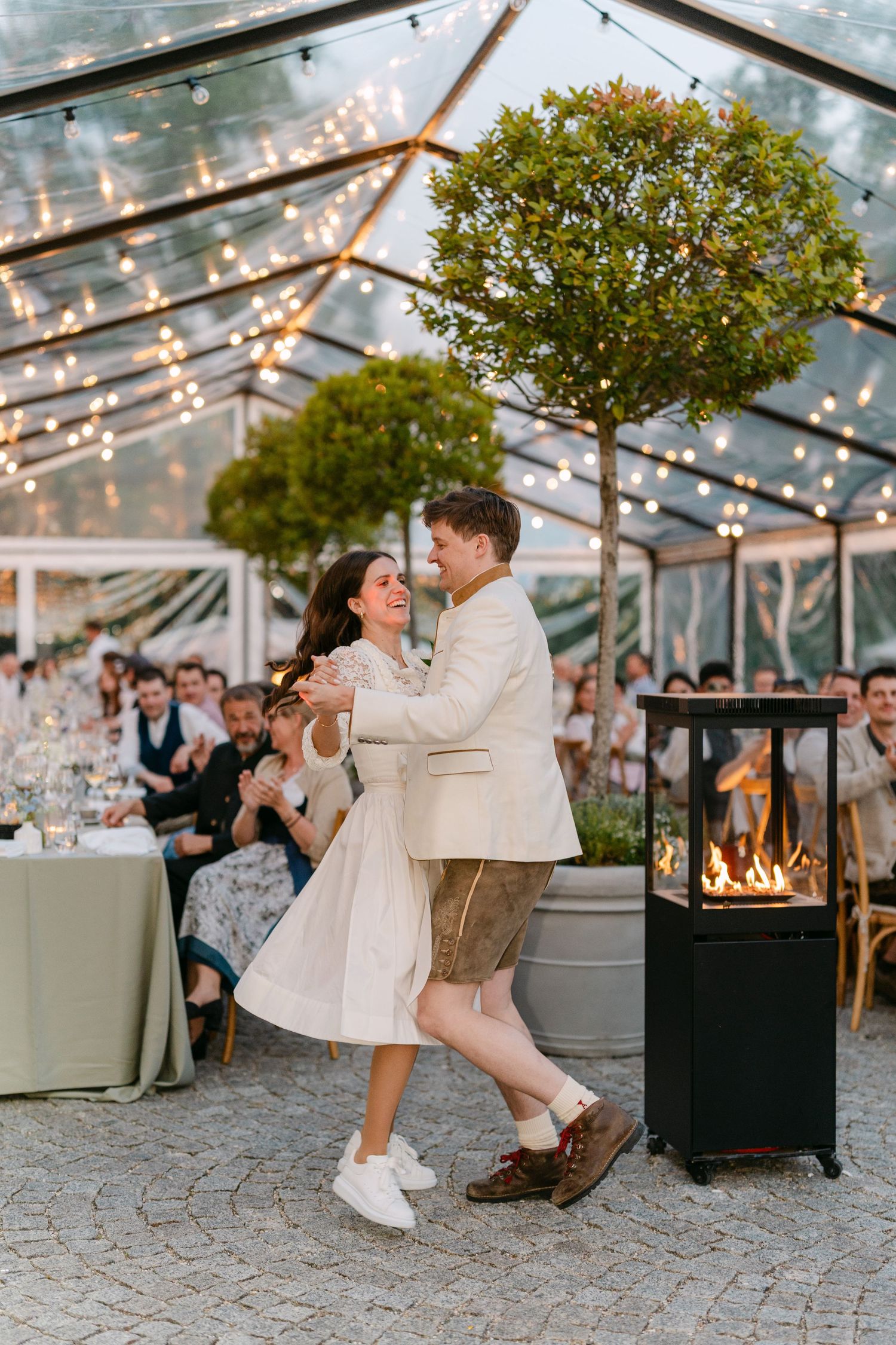 Romantic evening celebration under clear tent with string lights and outdoor fireplace.