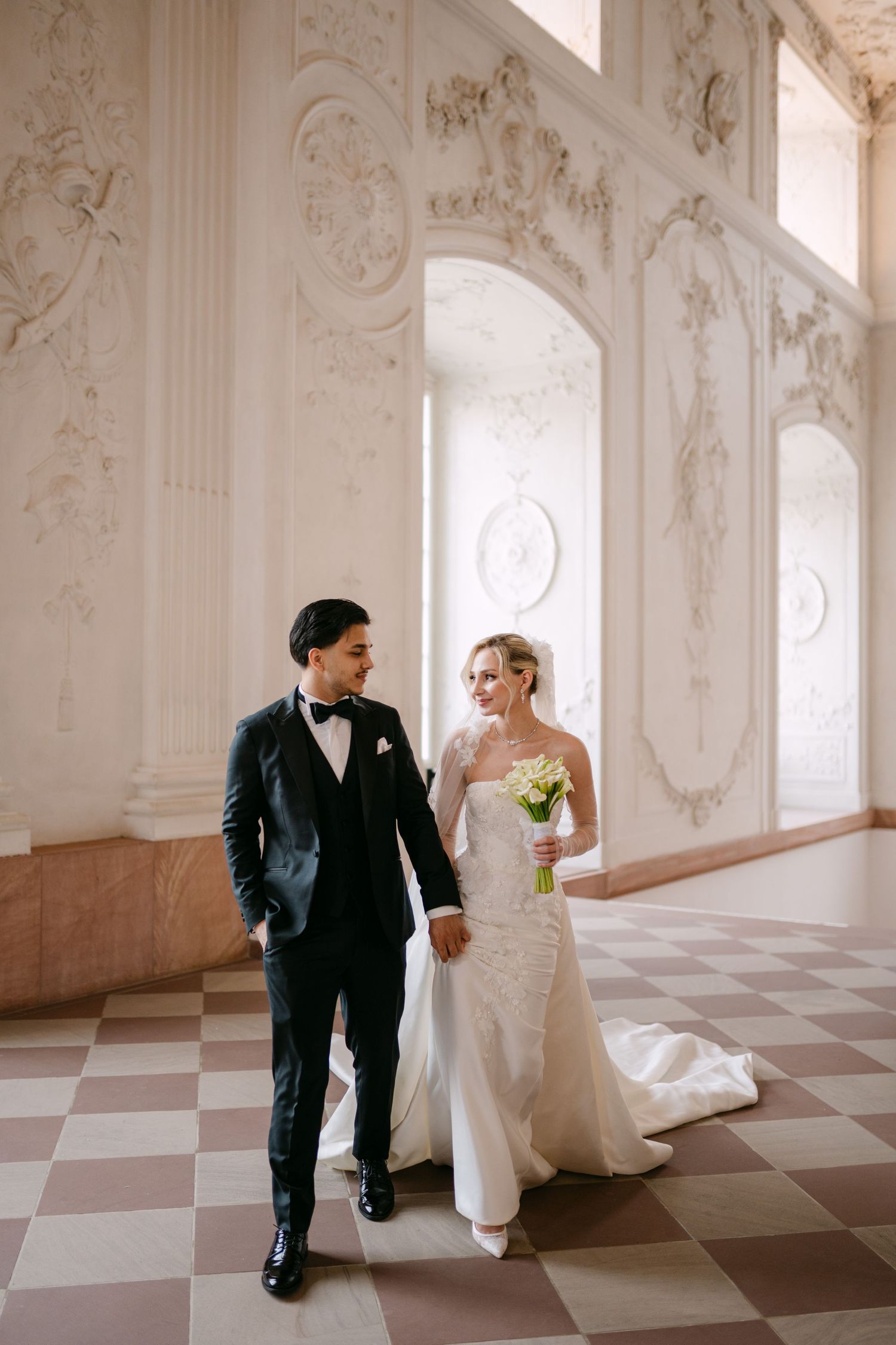 Wedding couple poses playfully in an ornate white ballroom with checkerboard flooring.