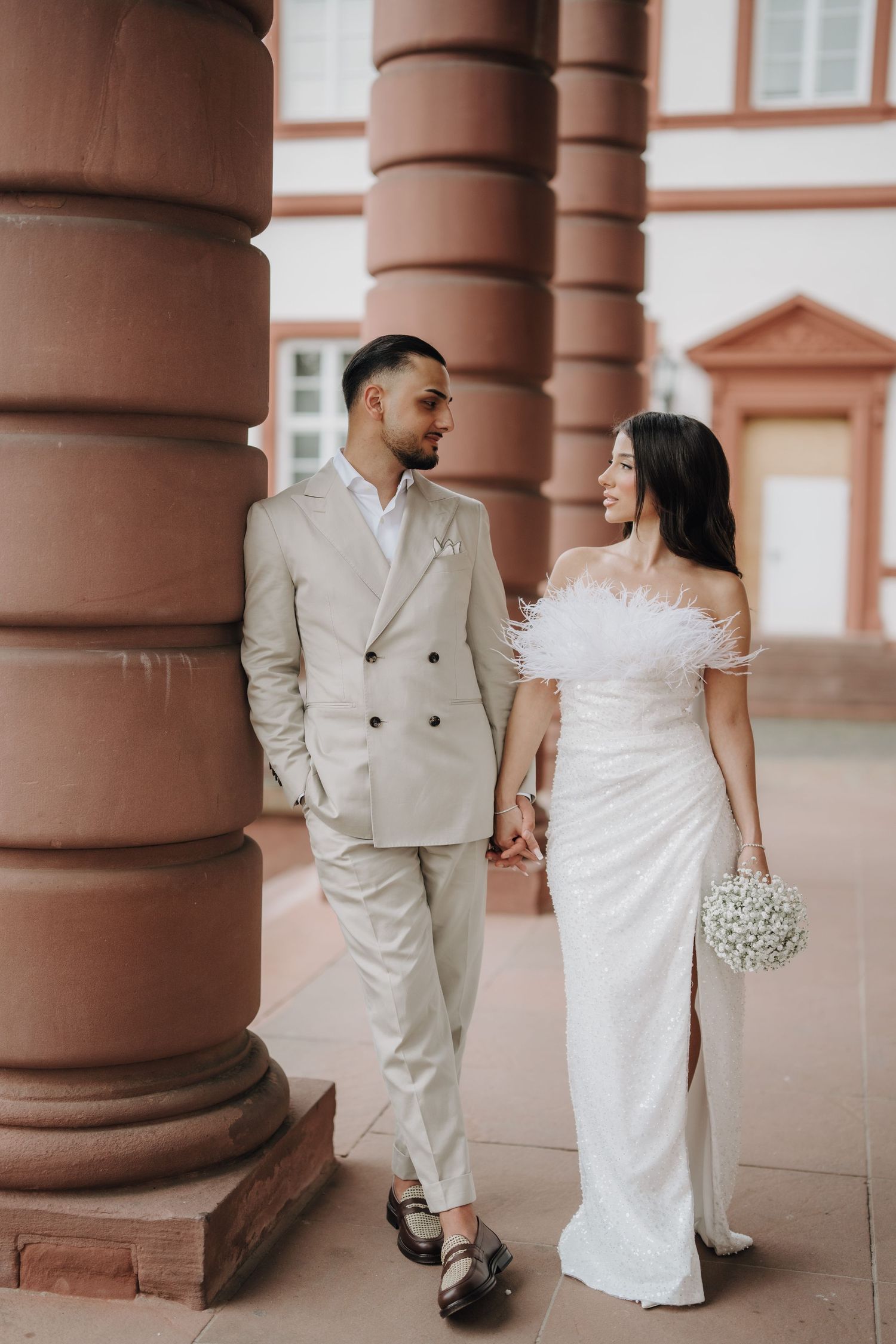 Elegant wedding couple poses between ornate terracotta columns in a romantic architectural setting.