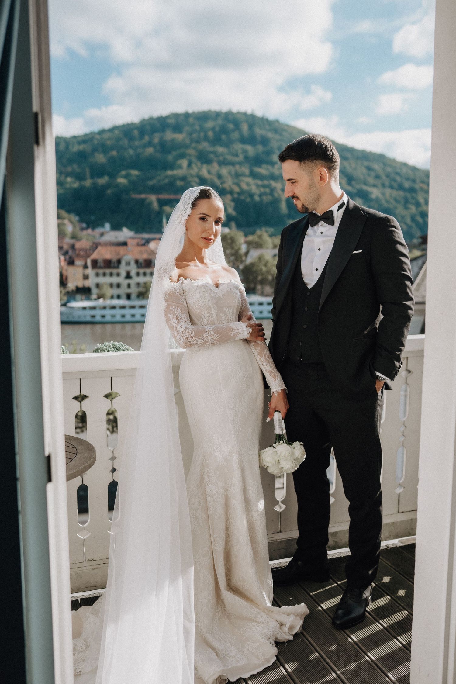 Elegant wedding couple on a balcony overlooking mountains and a river on a sunny day.