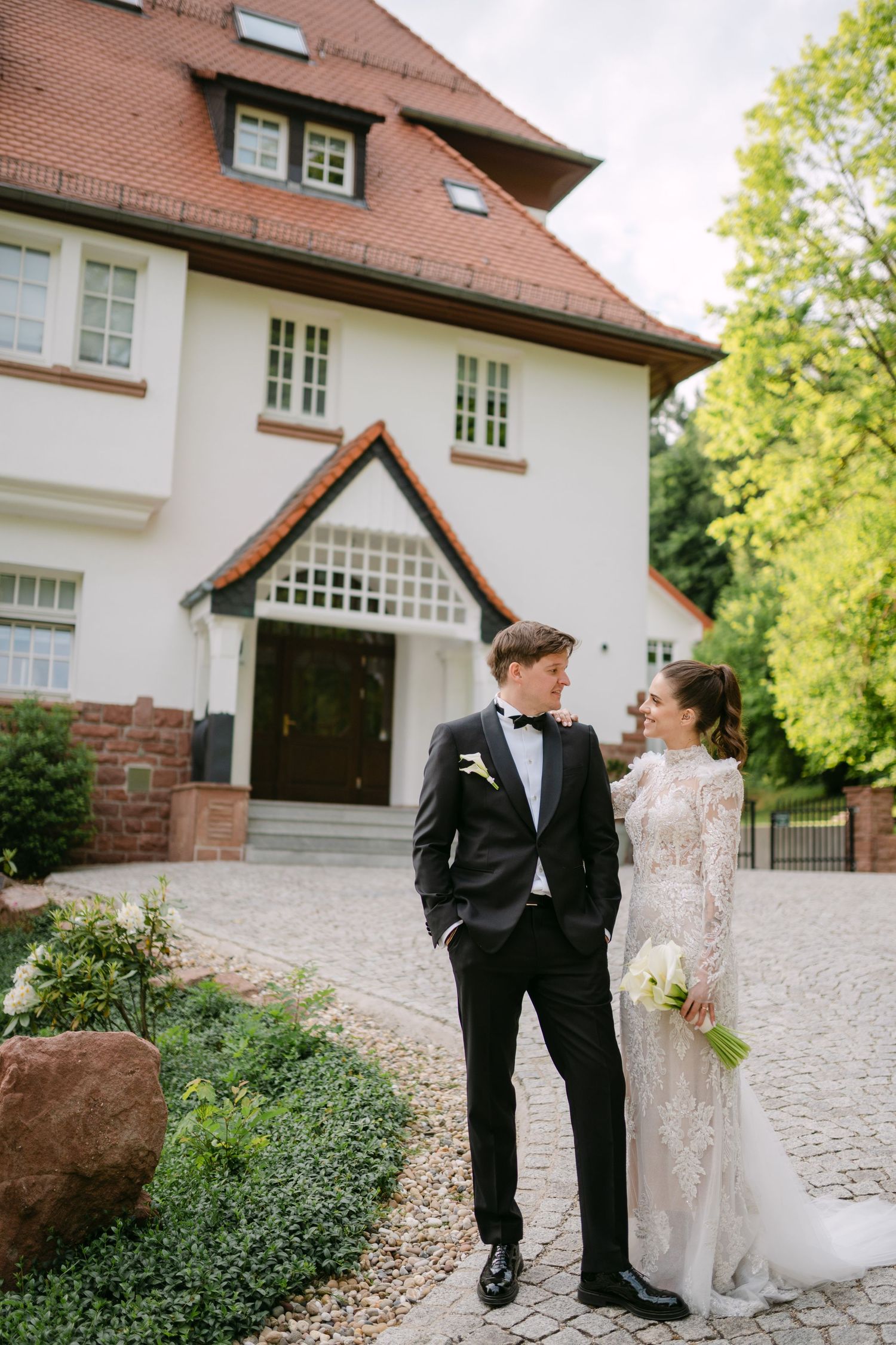 Wedding couple poses together outside a charming white house with brick accents and landscaped grounds.